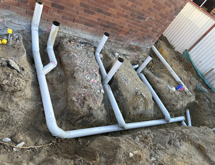 Gray PVC pipes in trenches near a brick wall, ready for plumbing installation.