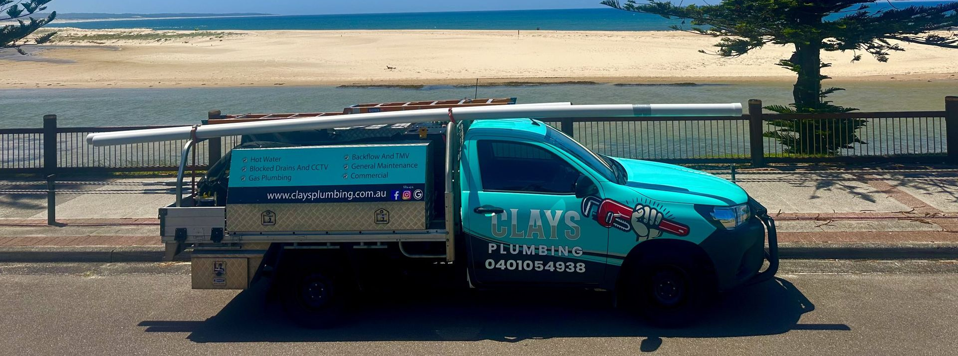 A teal plumbing truck parked near a beach. It has pipes on top and a logo on the side.