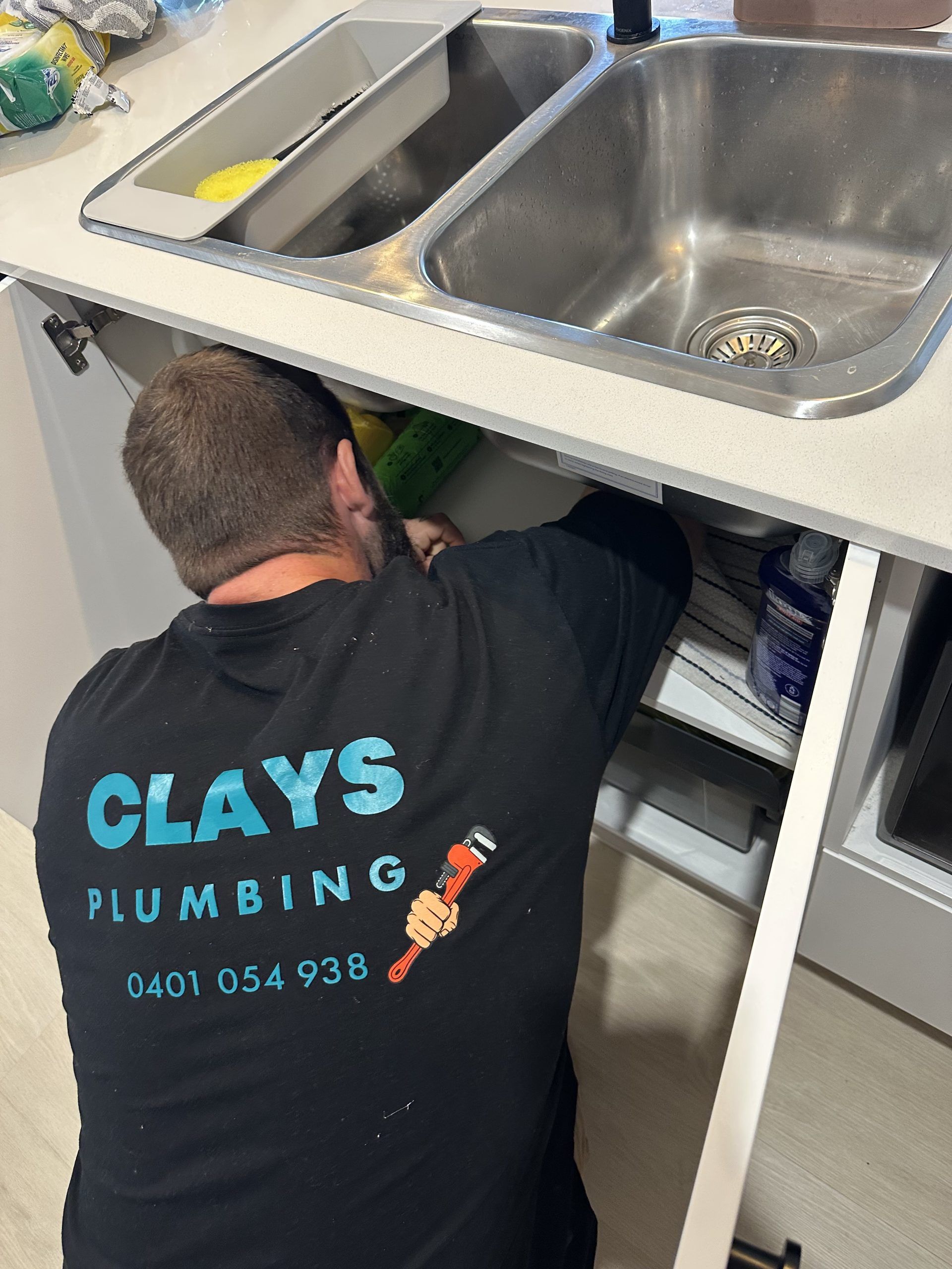 Plumber in black shirt works under a kitchen sink. Text on shirt: Clay’s Plumbing, phone number.