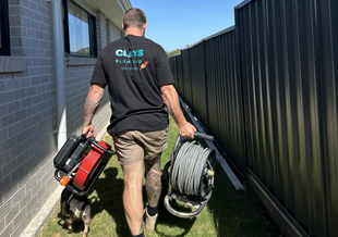Man carrying equipment, walking along a building with a black fence. Dog is at his feet.