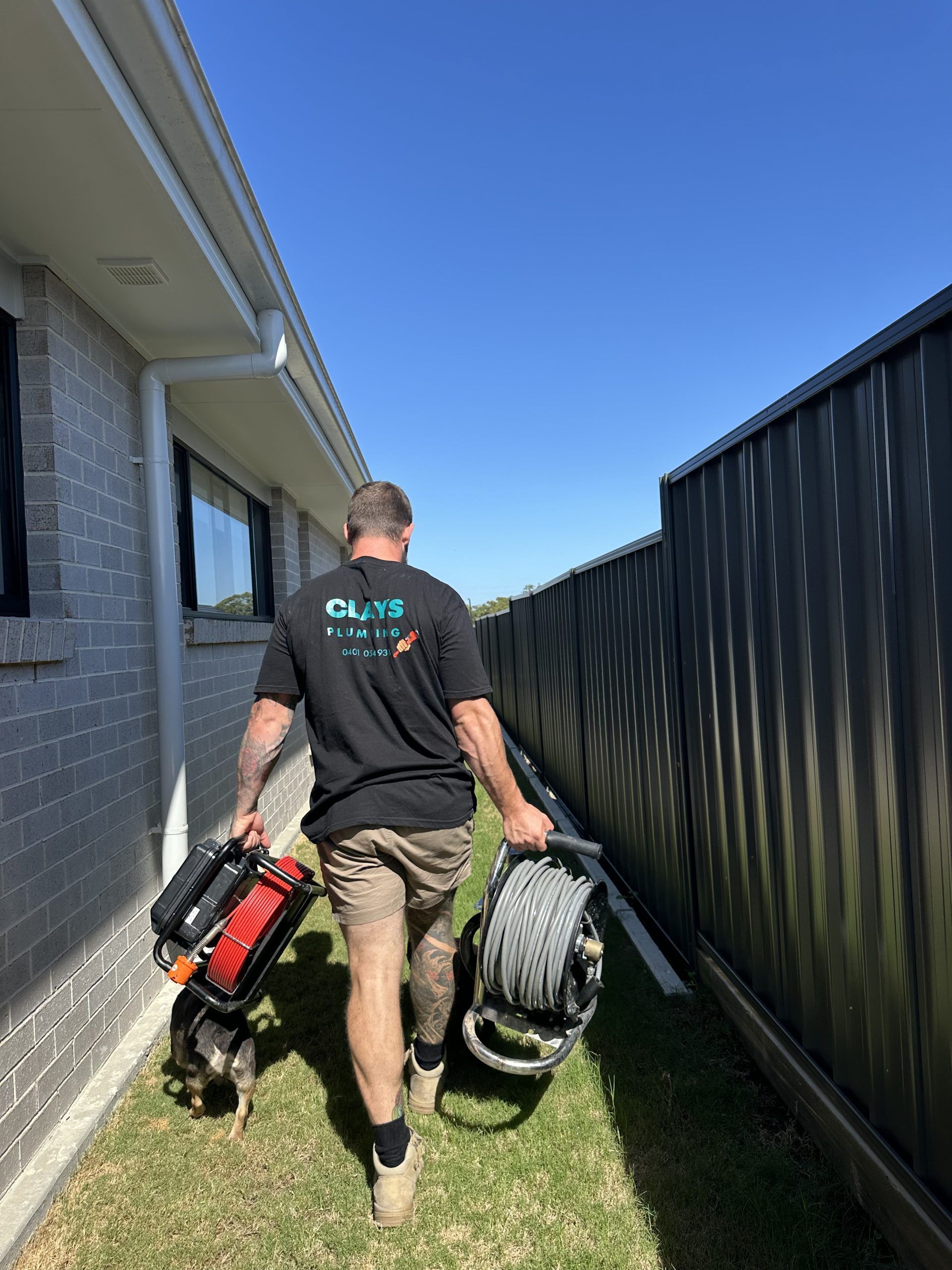 Man walking with tools and a dog down a narrow yard path, sunny day.