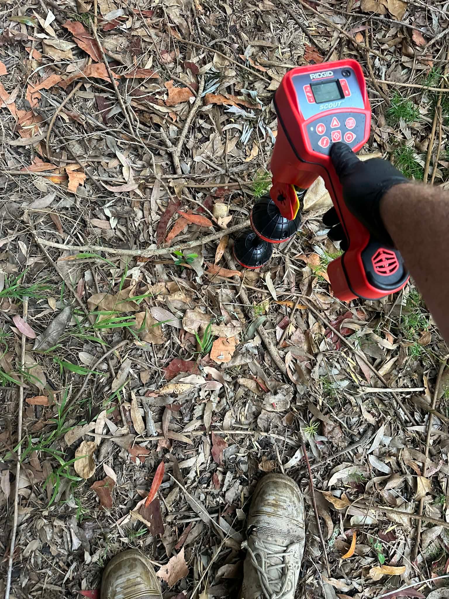 Person using a red handheld metal detector over leaf-covered ground, with boots visible below.