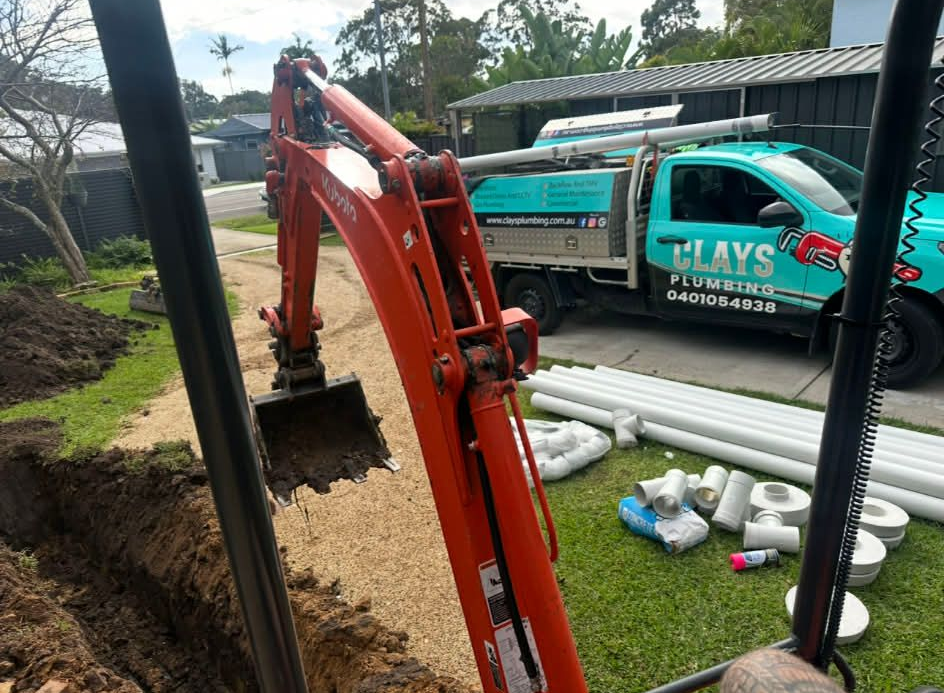 Red excavator beside a teal work truck at a roadside construction site with pipes on the grass