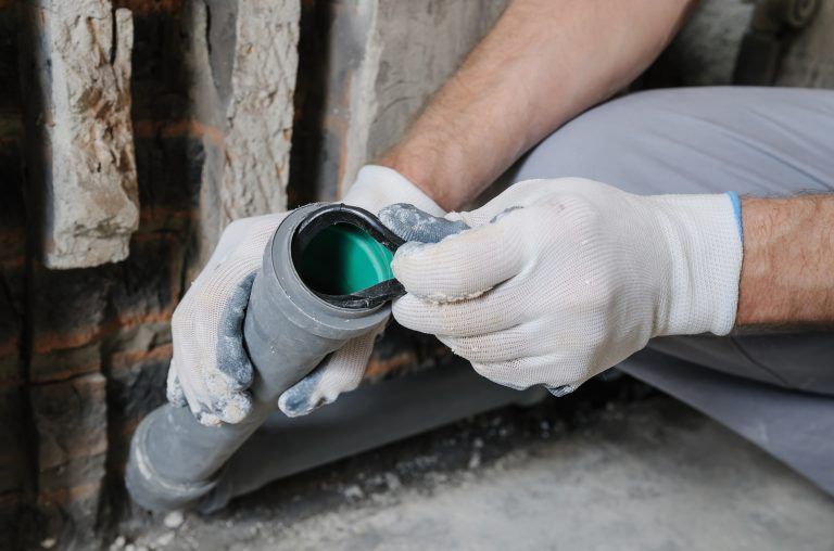 Person in gloves seals a gray pipe with a green cap, near a brick wall, indoors.