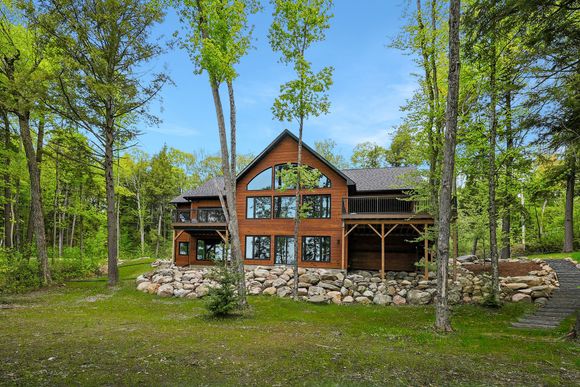 Wooden cabin with stone foundation in a sunny forest clearing, surrounded by tall green trees.