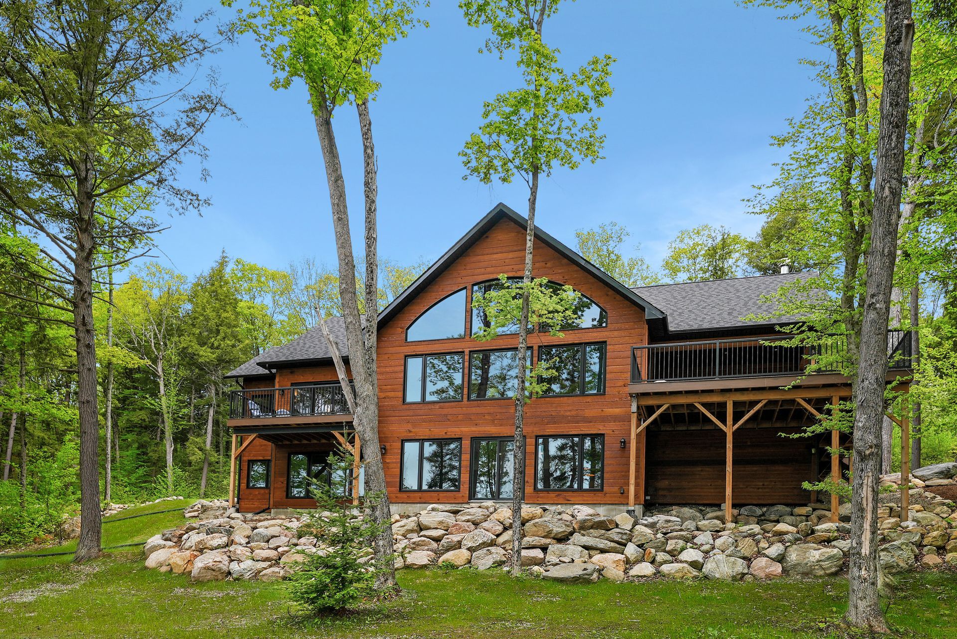 Rustic cabin with large windows and a stone foundation surrounded by green trees under a blue sky