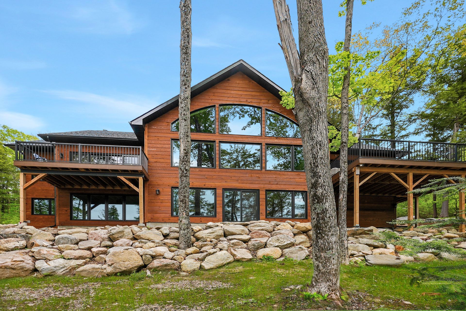 Large wooden cabin with stone base, tall windows, and wraparound deck among trees by a grassy yard