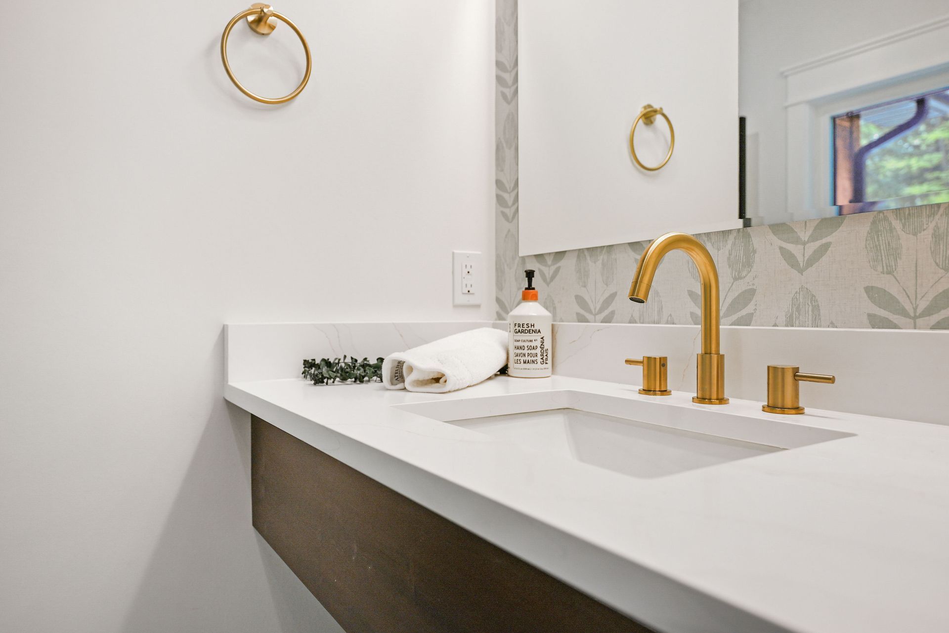 Modern bathroom vanity with white countertop, gold faucet, and round wall mirror lighted by a window.