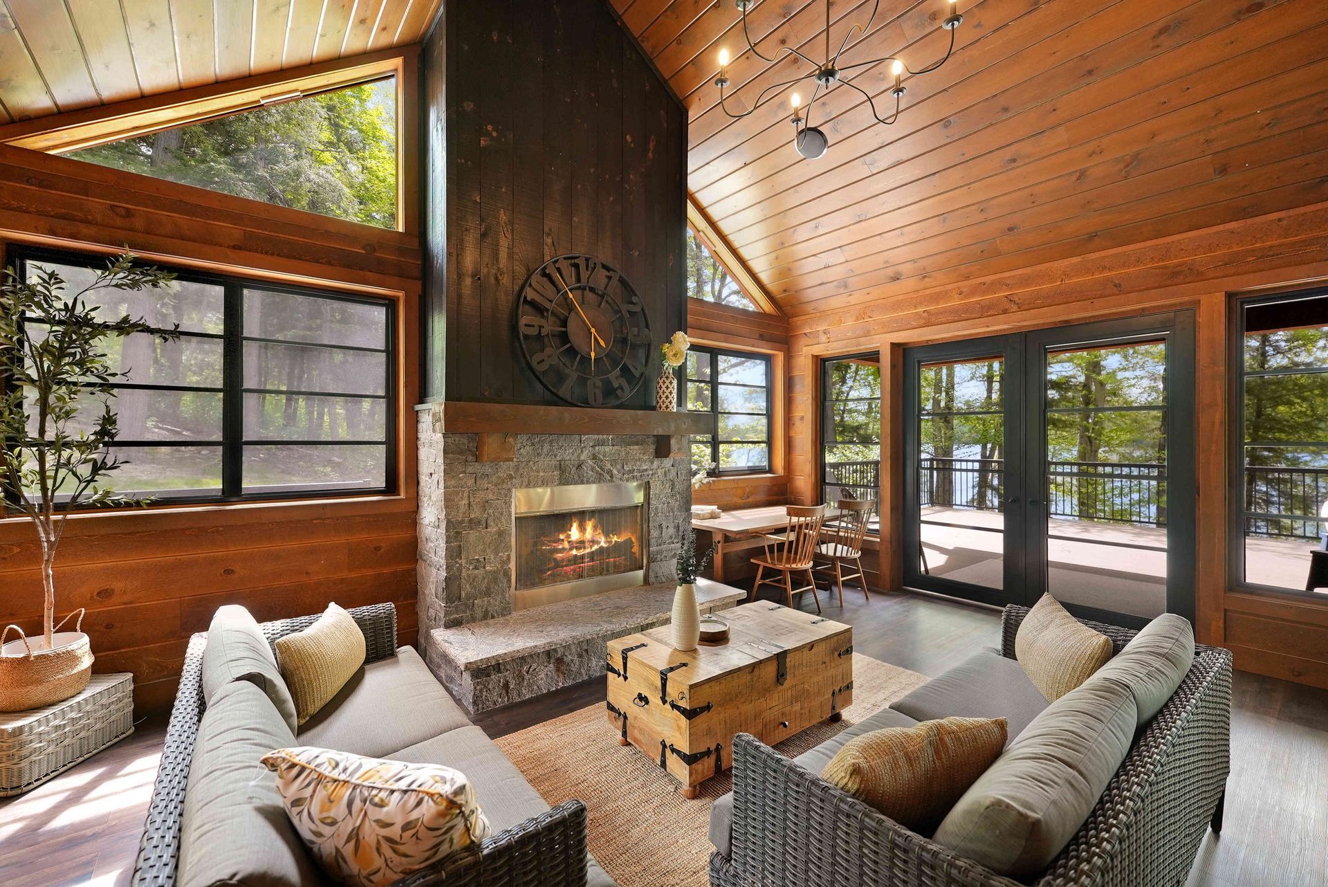 Cozy cabin living room with stone fireplace, wood-paneled ceiling, and large windows overlooking trees