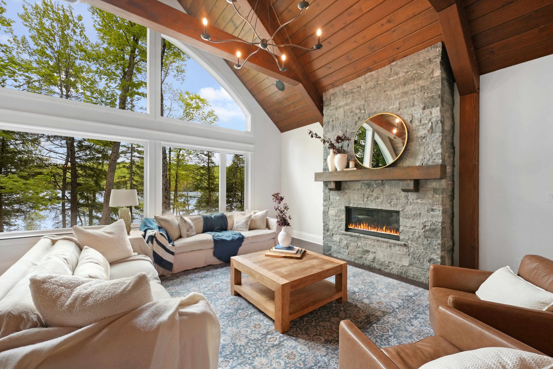 Bright living room with stone fireplace, large windows, wood ceiling, and beige seating around a coffee table