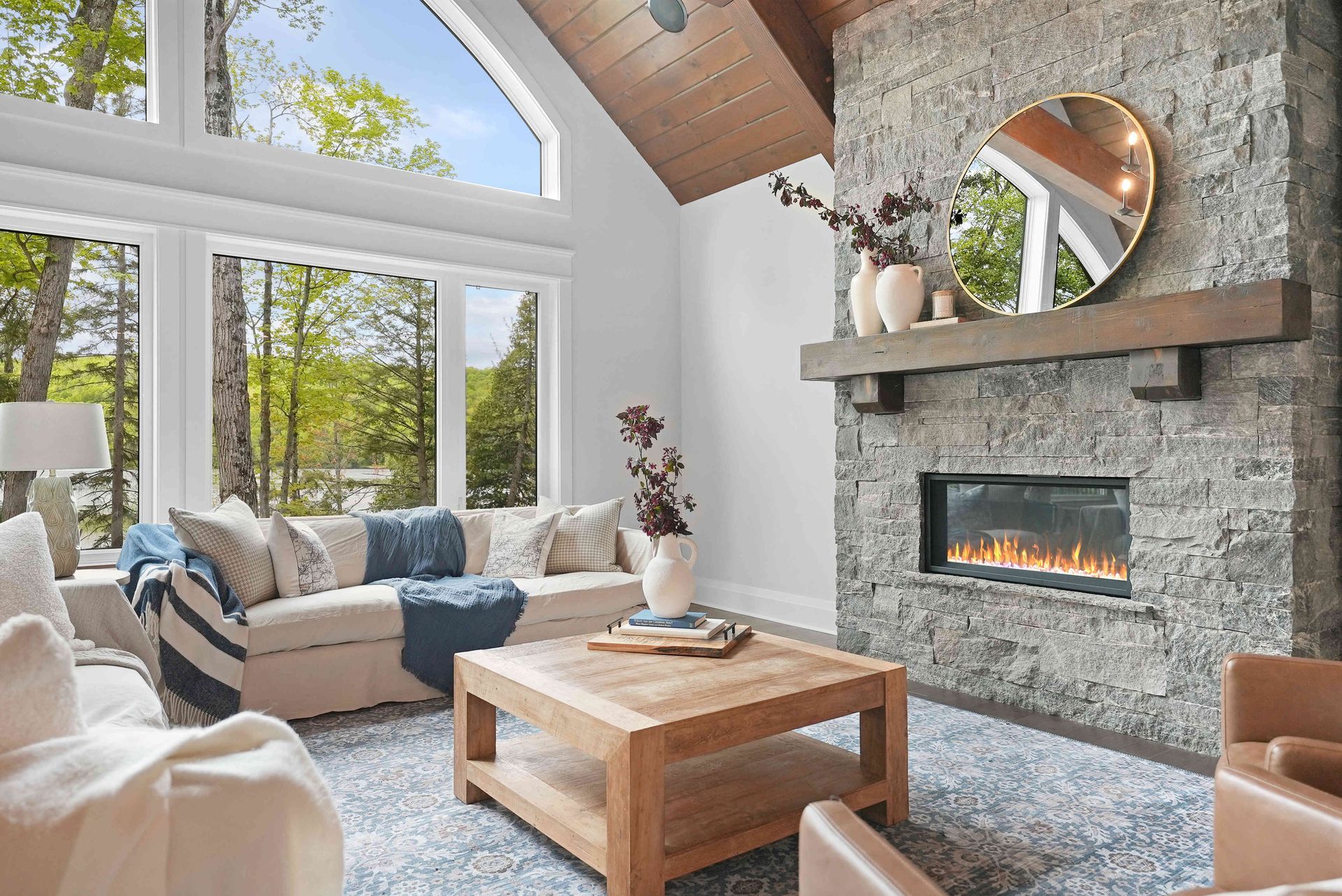 Bright living room with stone fireplace, large windows, beige sofa, and wooden coffee table