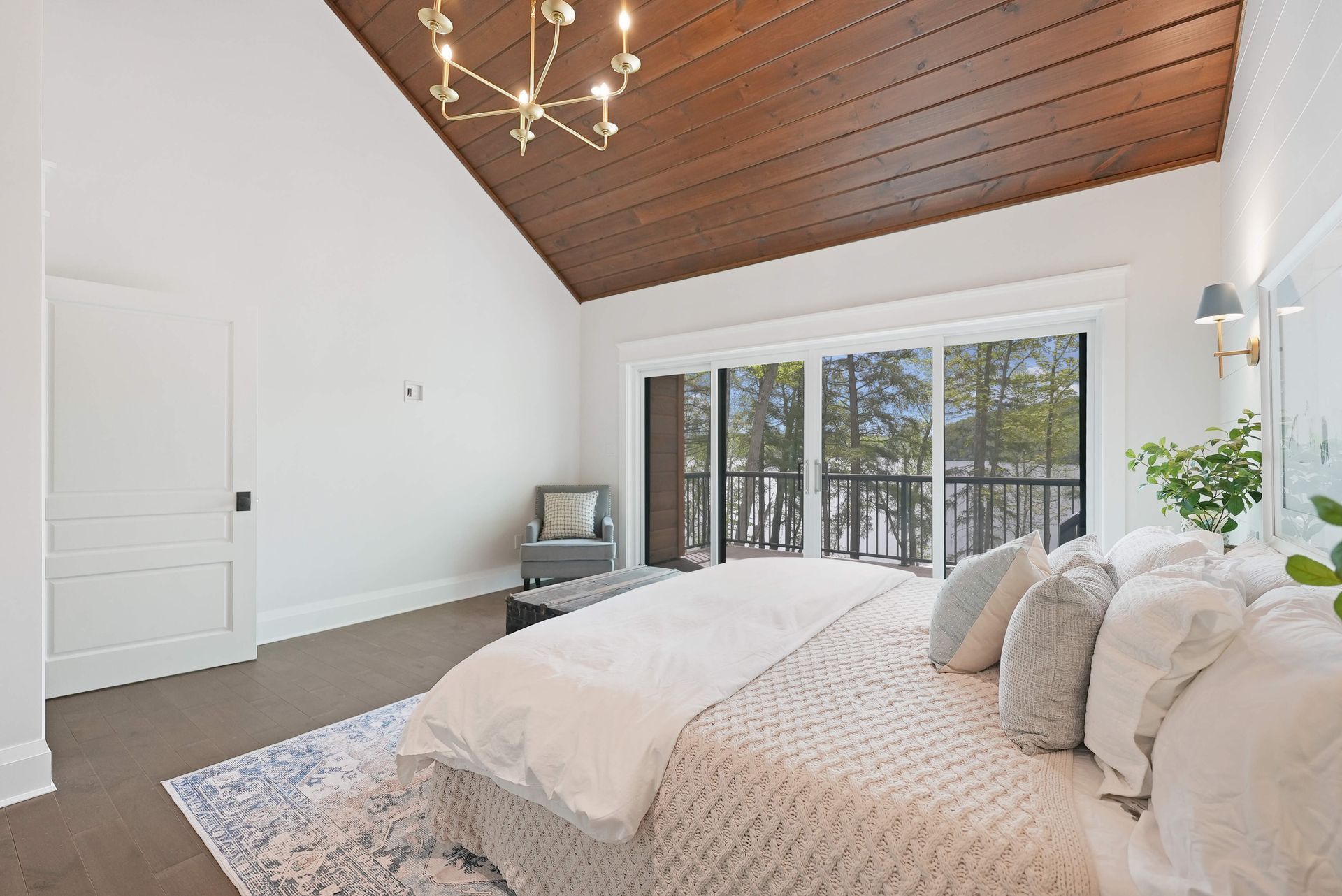 Bright bedroom with a wood-paneled vaulted ceiling, white bedding, and large windows overlooking trees.