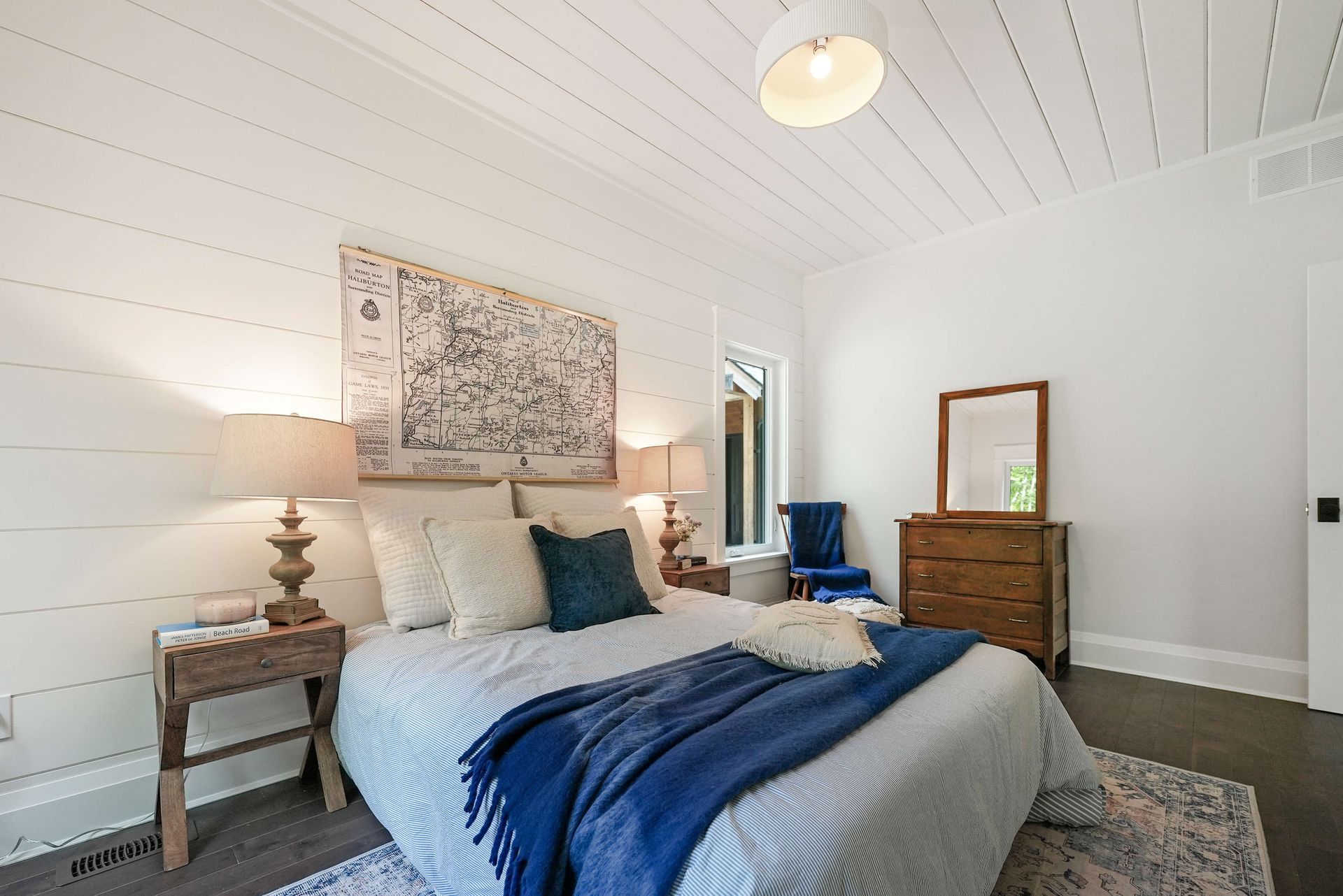 Bright bedroom with blue bedding, wooden dresser, bedside lamps, and a patterned wall hanging.