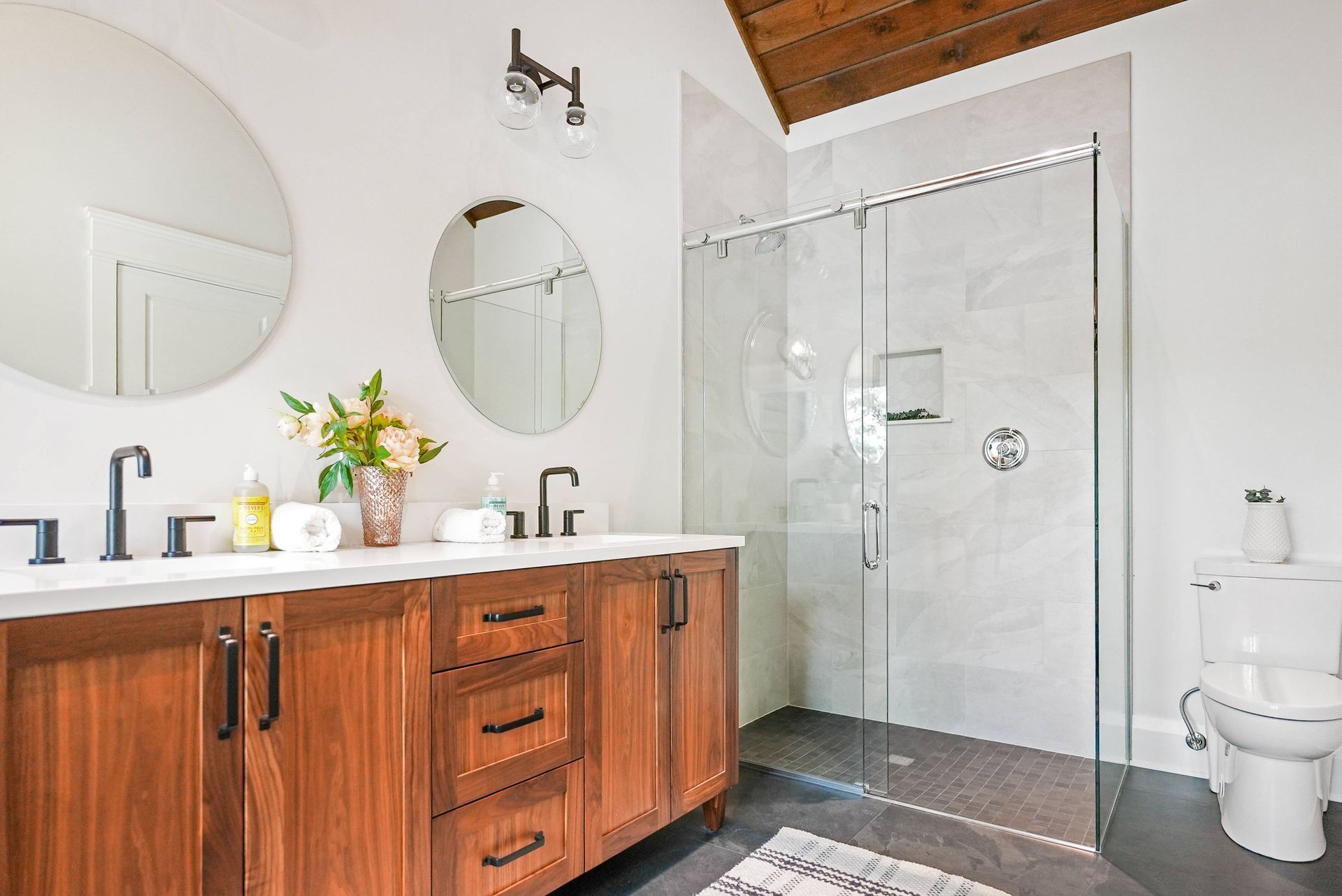 Modern bathroom with wooden vanity, round mirrors, glass shower, and a toilet under a vaulted wood ceiling