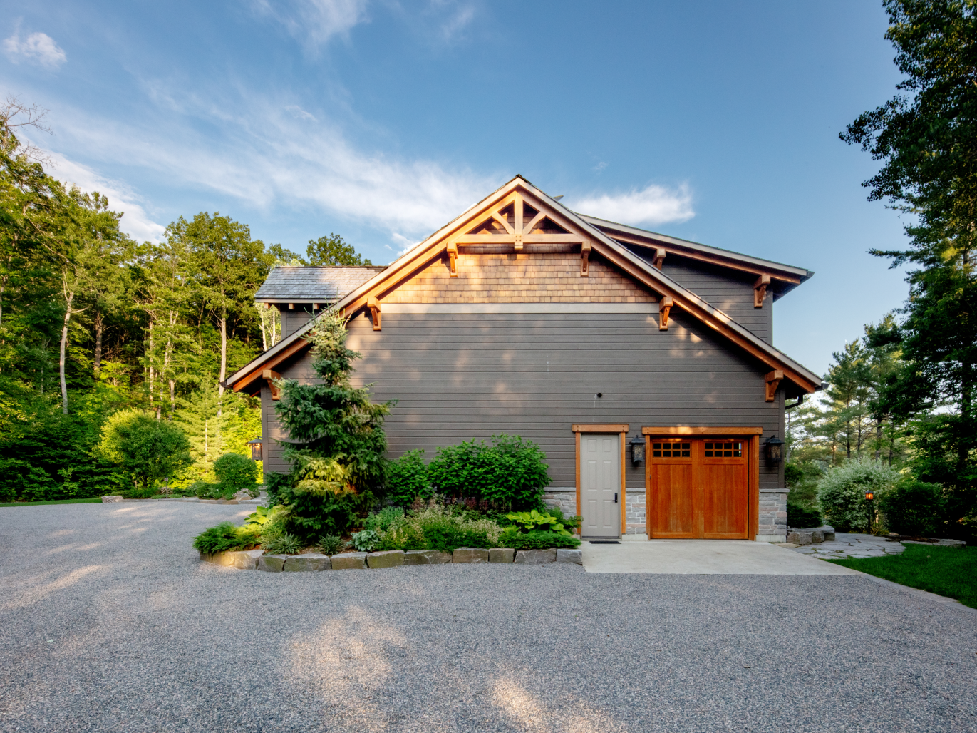 A large house with a wooden garage door is surrounded by trees and gravel.