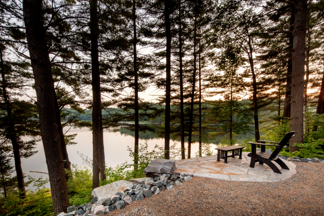 A patio with chairs and a table overlooking a lake in the woods.