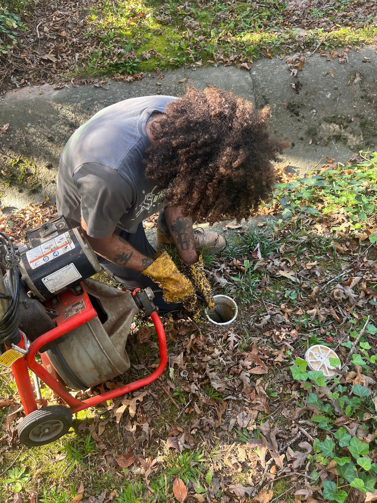 Person using a drain cleaning machine near a drainpipe. Outdoors, working on the ground.