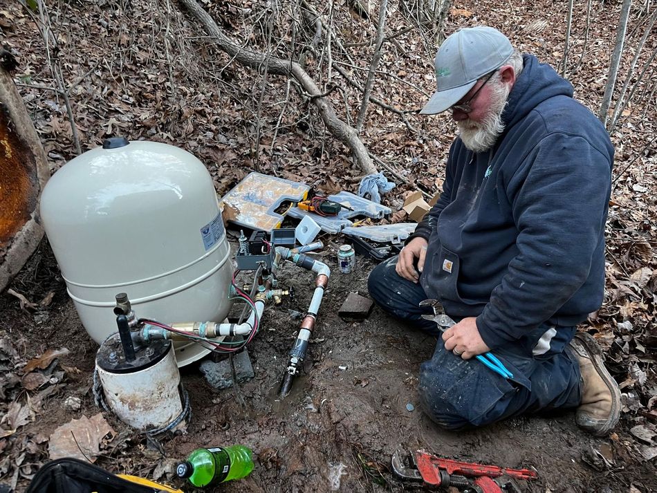 A man is kneeling down in the dirt working on a water pump.