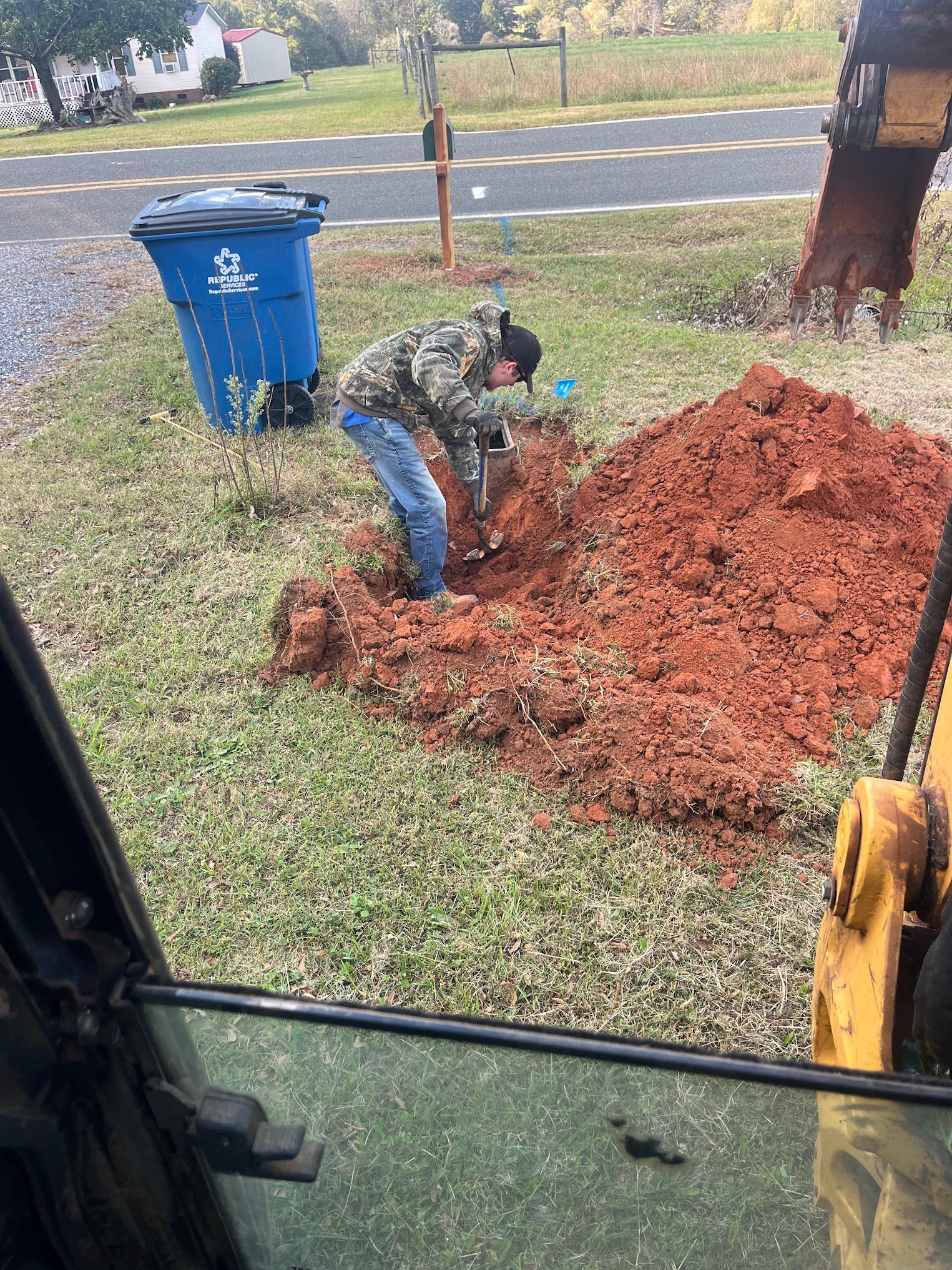 A man is digging a hole in the ground in front of a blue trash can.