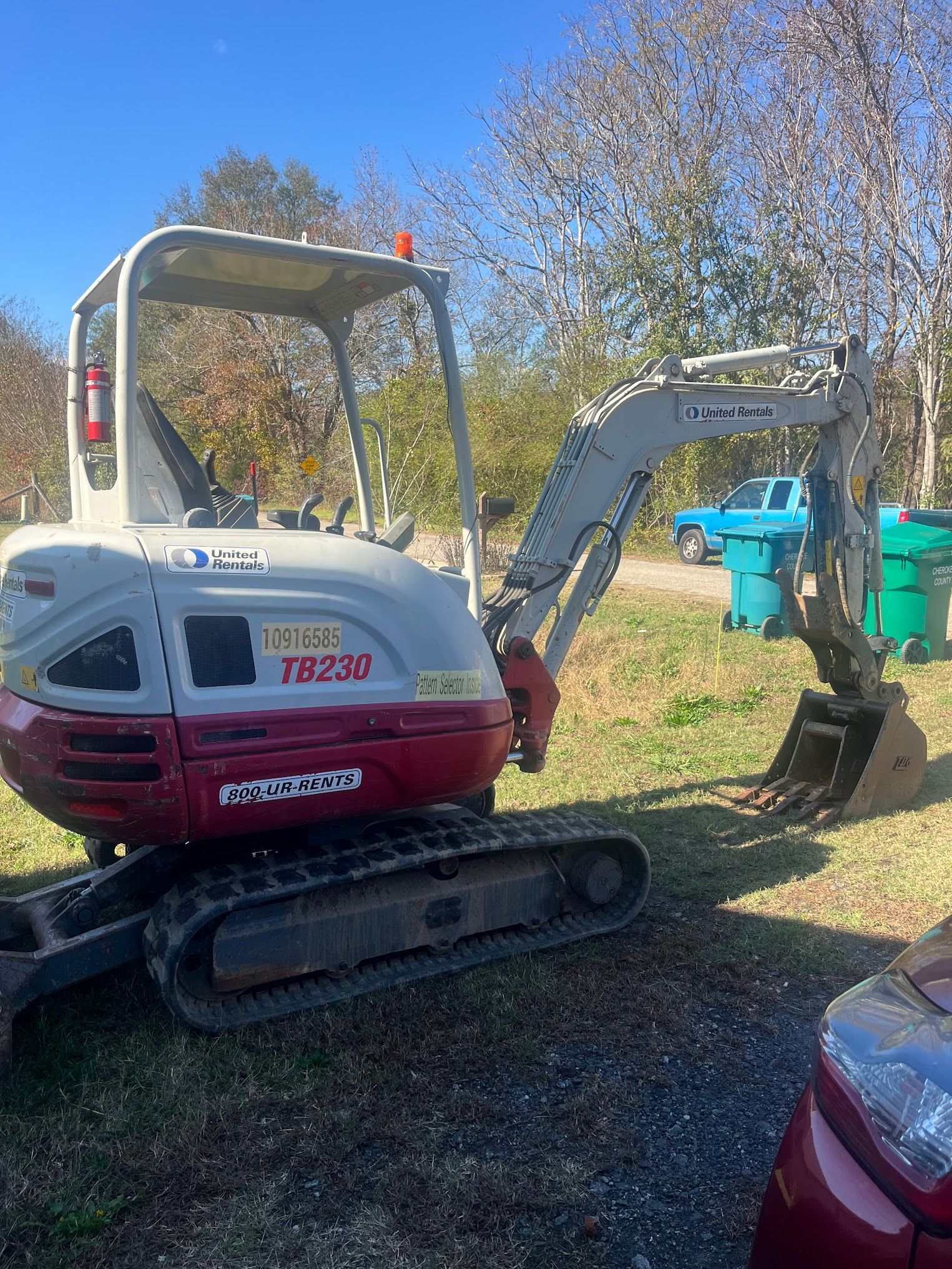 A small excavator is parked in a grassy field next to a red car.