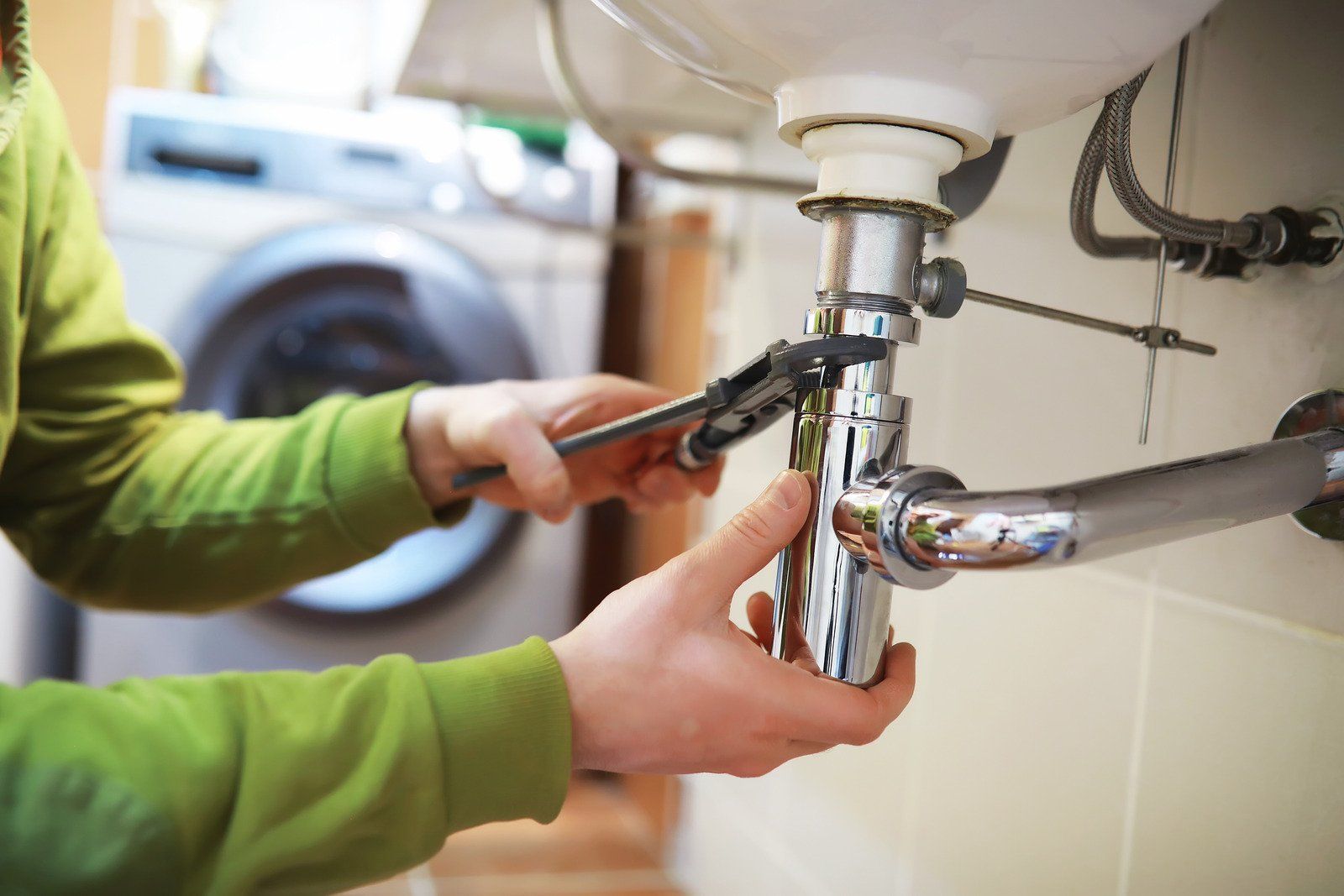 Person using a wrench to repair plumbing under a sink. A washing machine is visible in the background.