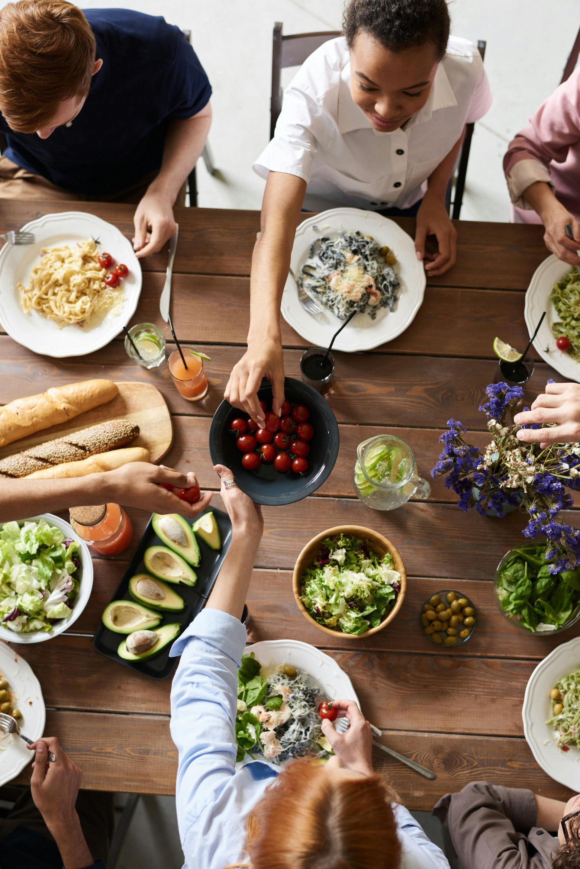People reaching for food at a wooden table set with dishes, salads, tomatoes, and drinks.