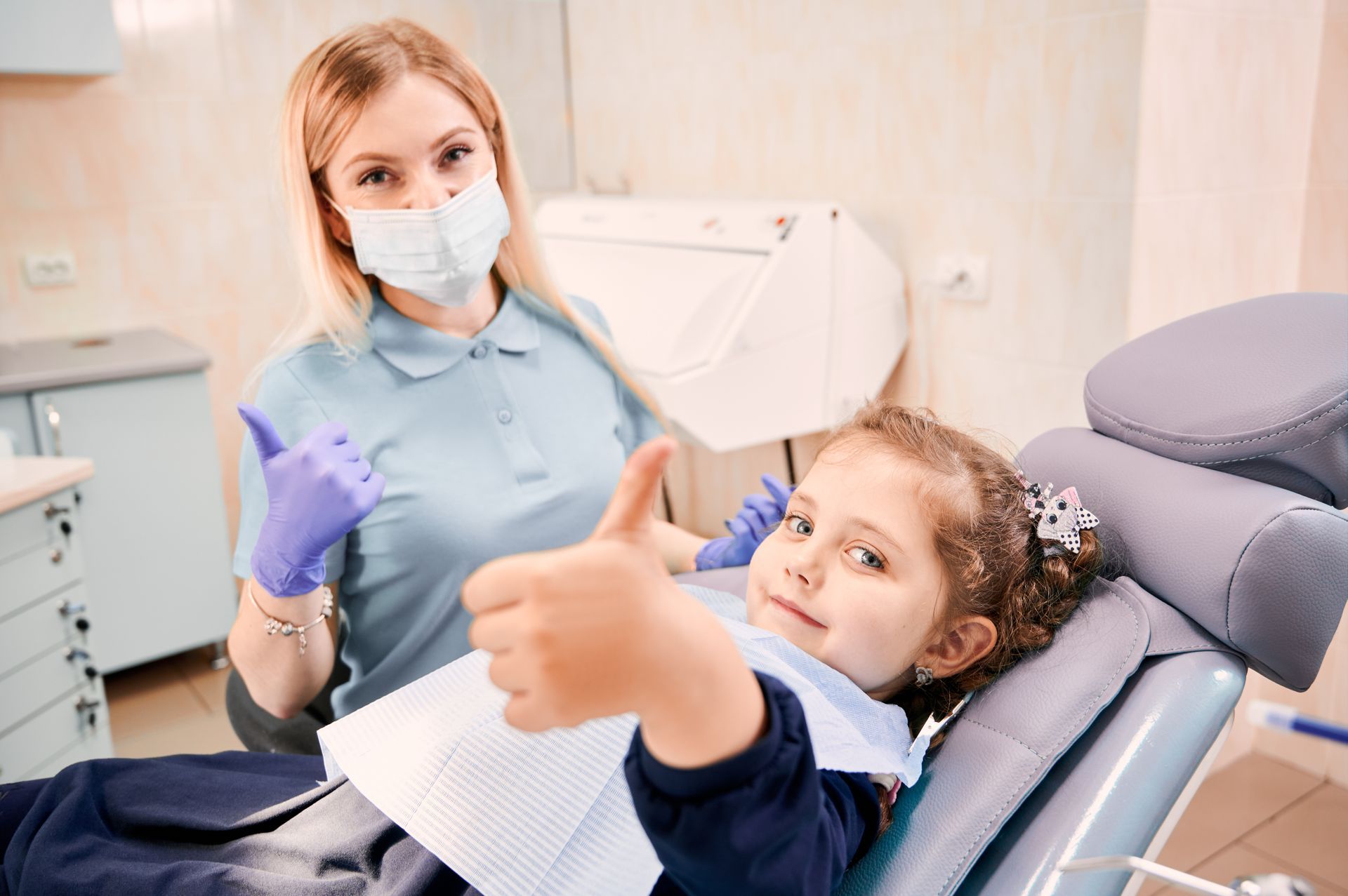 A little girl is giving a thumbs up while sitting in a dental chair.