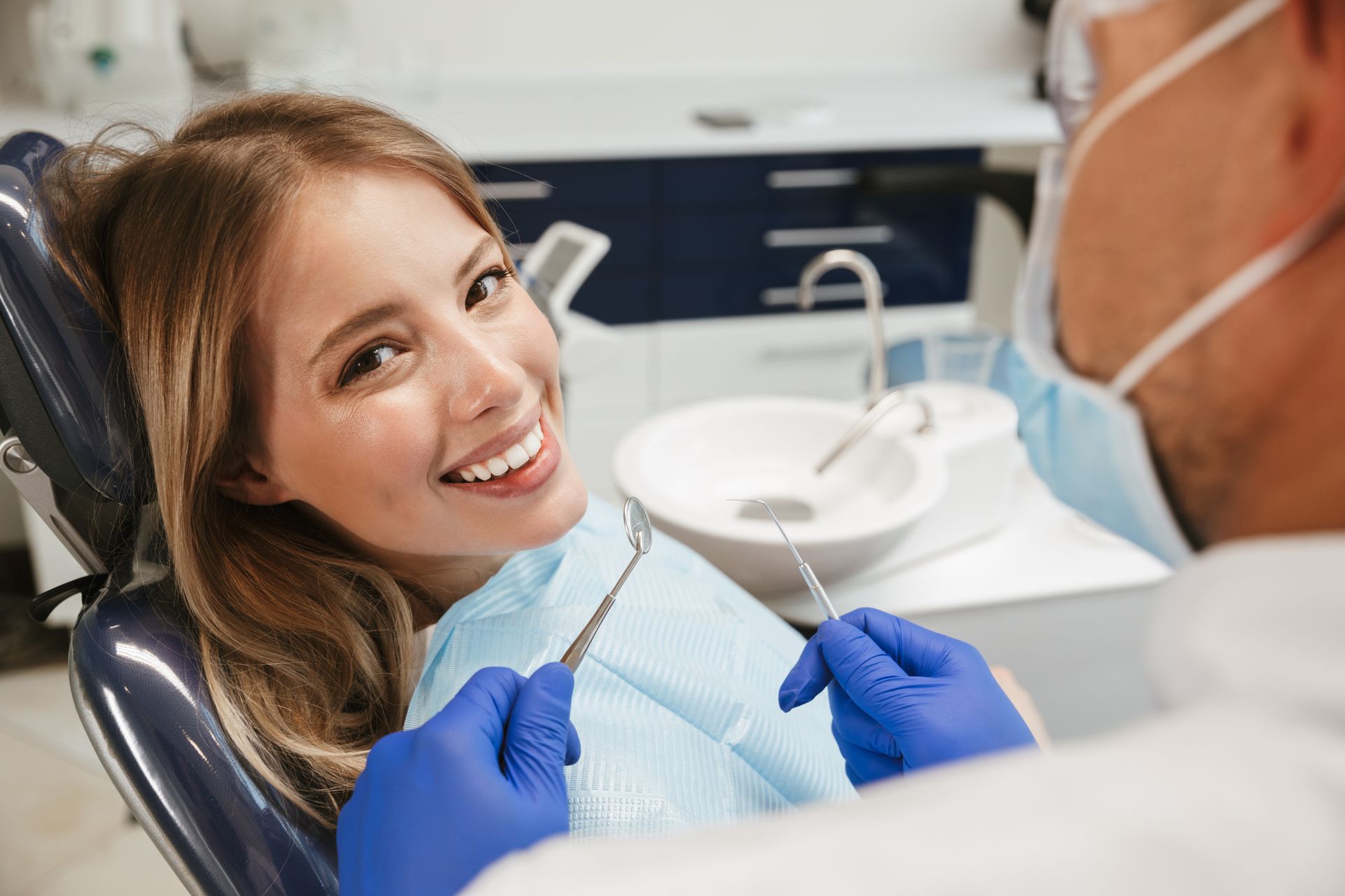 A woman is smiling while sitting in a dental chair while a dentist examines her teeth.