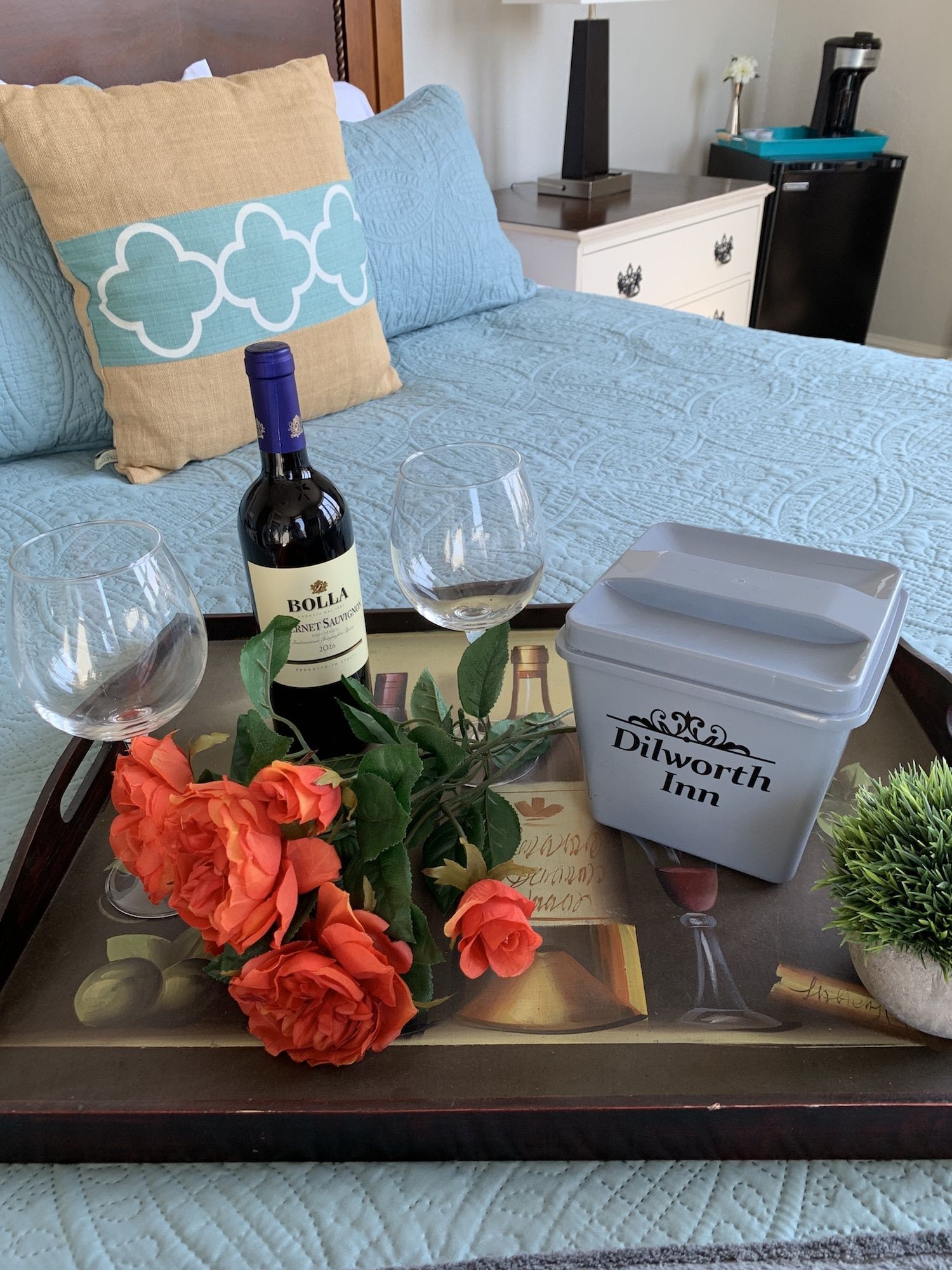 A tray with a bottle of wine , two glasses of wine , flowers and a bucket on a bed.