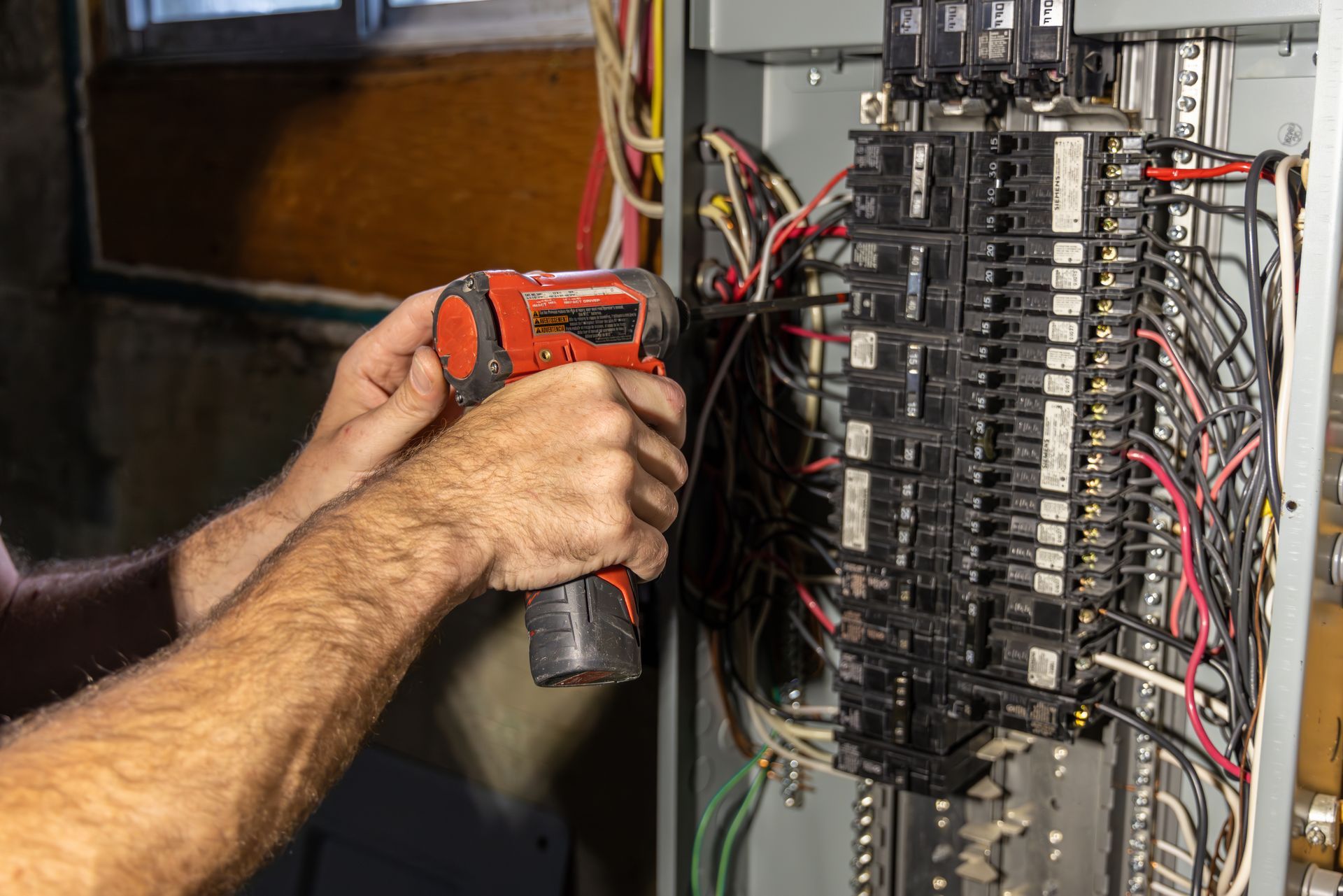 Person using a power tool on an electrical panel with wires; indoor setting.