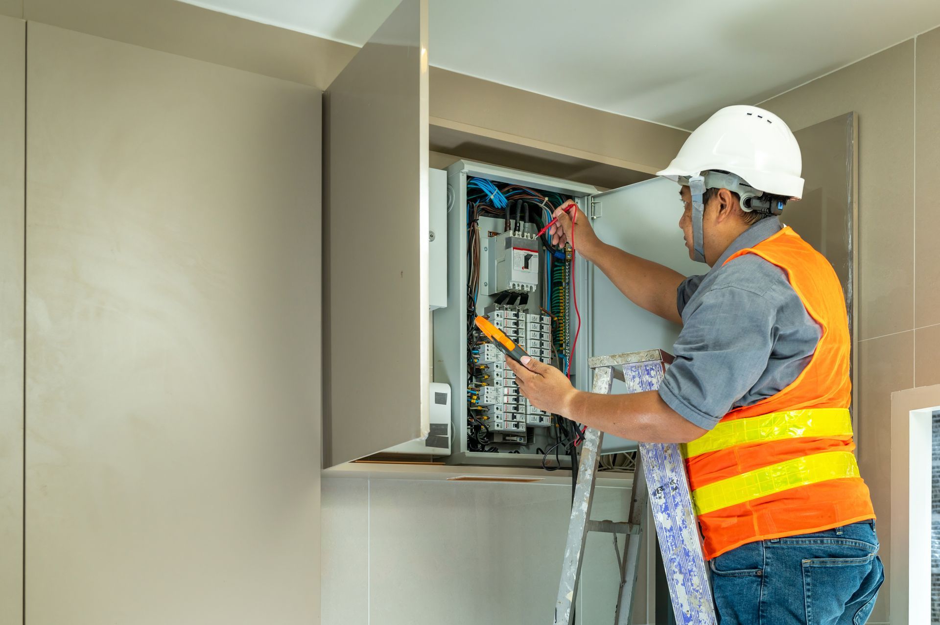 Electrician working on electrical panel, wearing a safety vest and helmet, standing on a ladder.