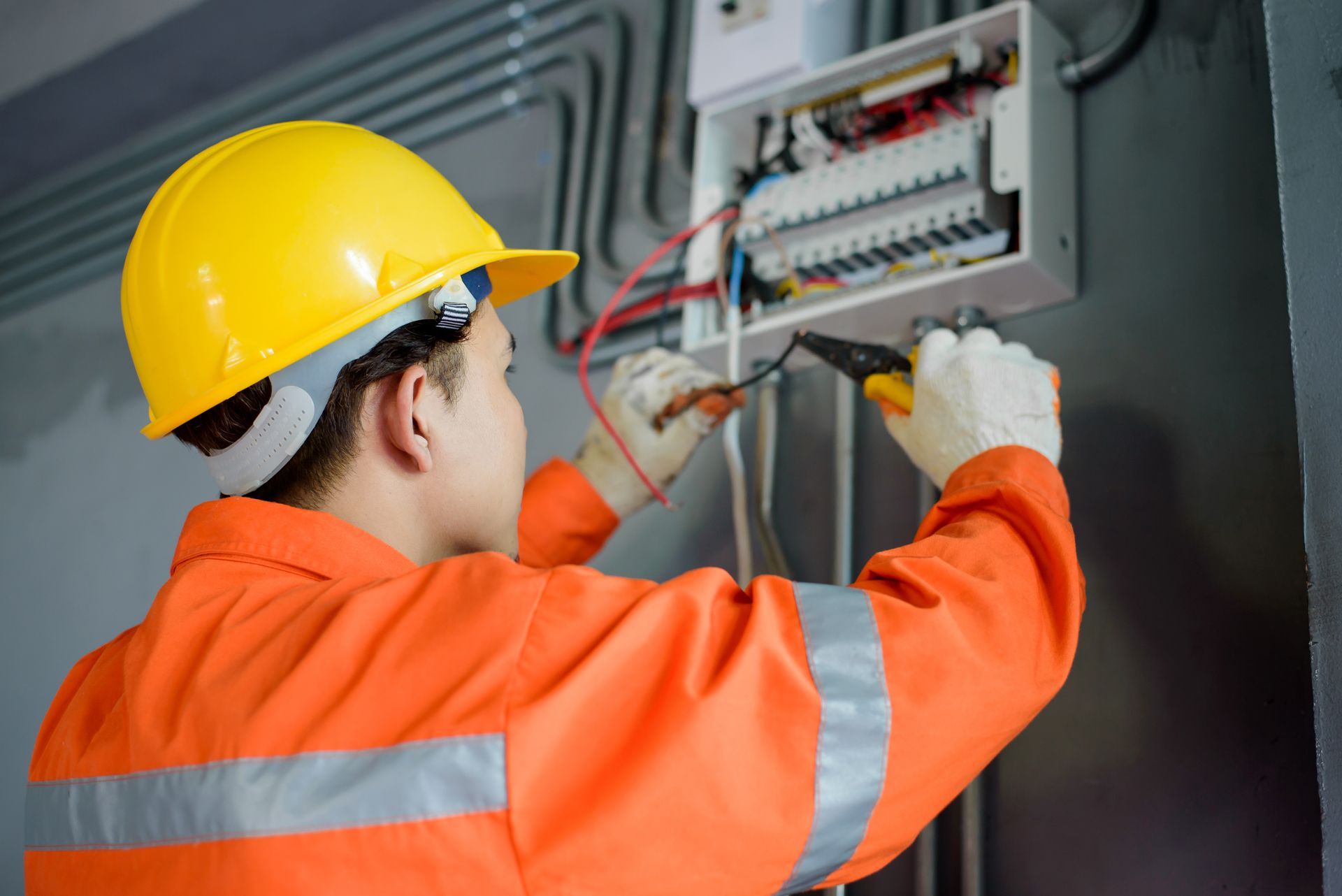 Electrician in orange jumpsuit and yellow hard hat working on an electrical panel.