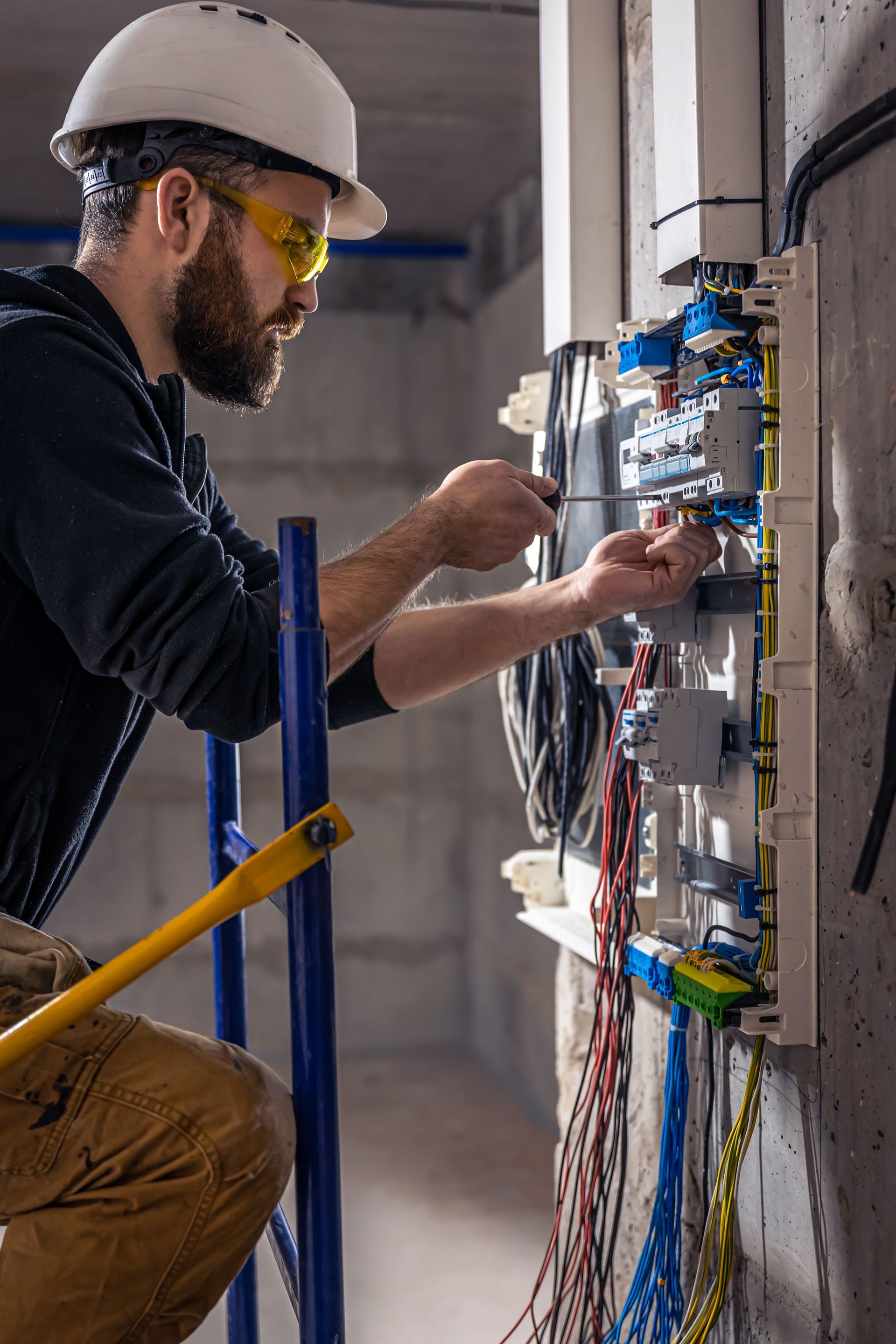 Electrician working on electrical panel, wearing safety glasses and a hard hat, standing on a ladder.