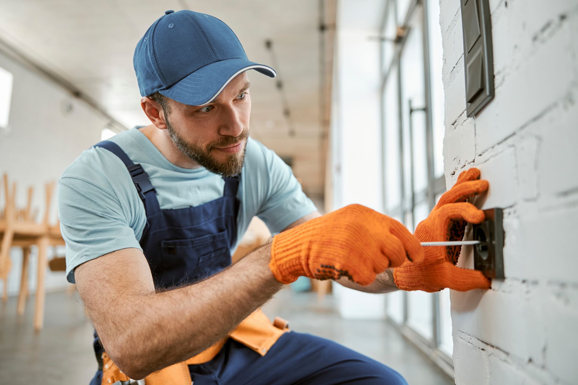 Electrician in orange gloves using a screwdriver on a wall switch. Brick wall, indoors.