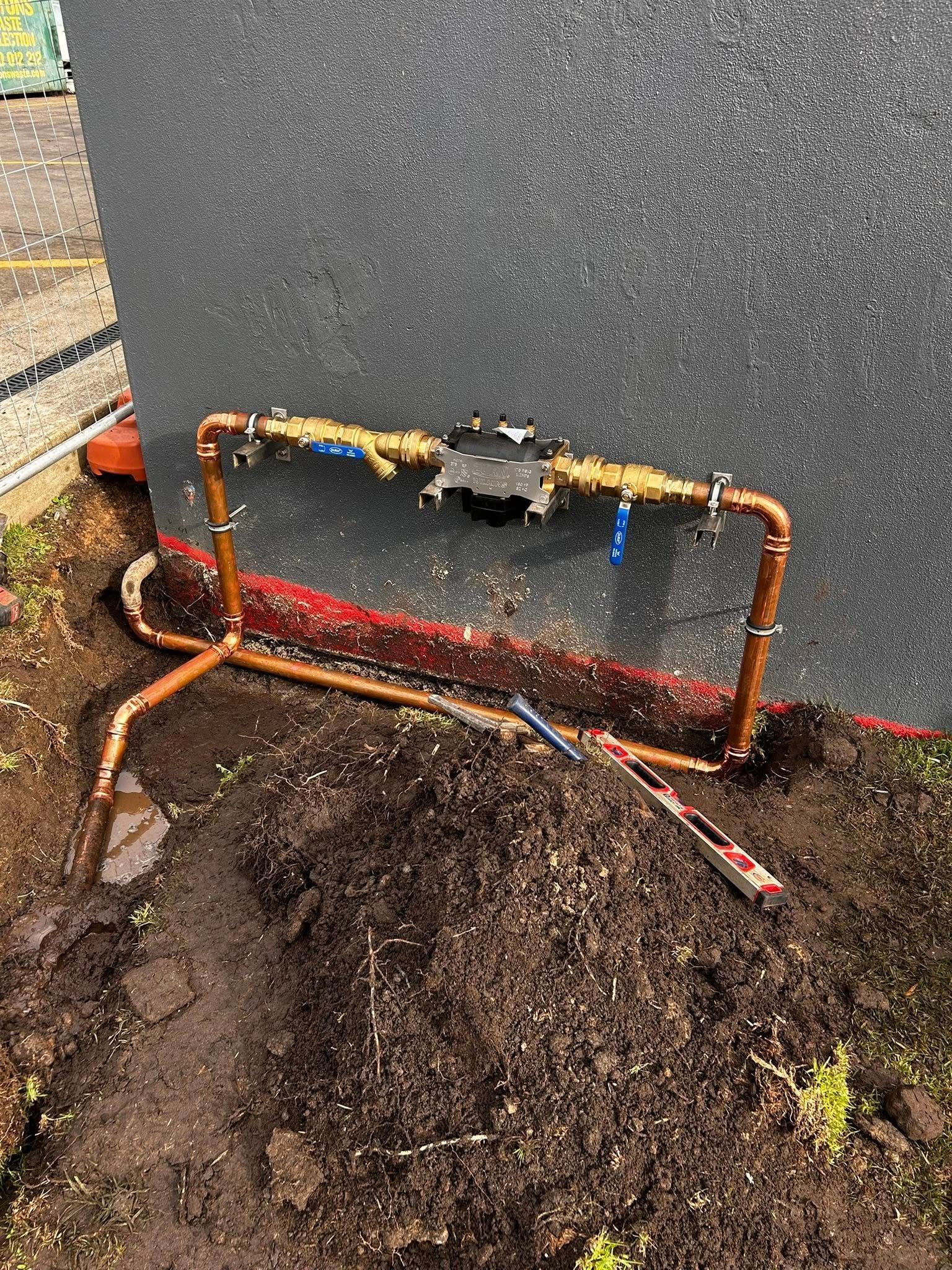 Copper pipes installed near a wall, in dirt. A black device is in the pipes. Blue and yellow valves are attached.
