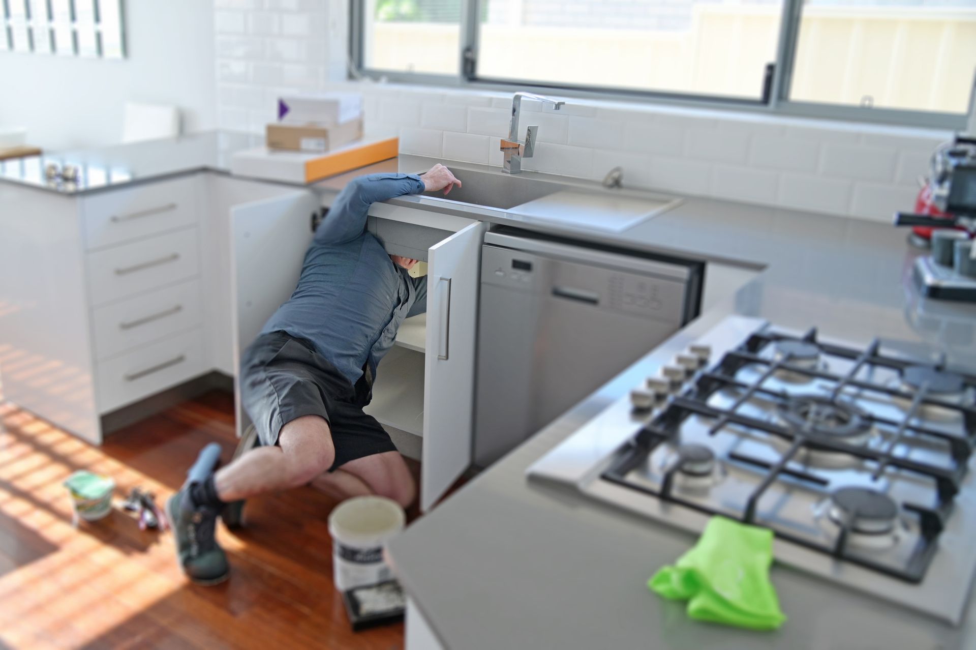 Plumber working under a kitchen sink in a modern kitchen with white cabinets, gray countertops, and a gas stove.