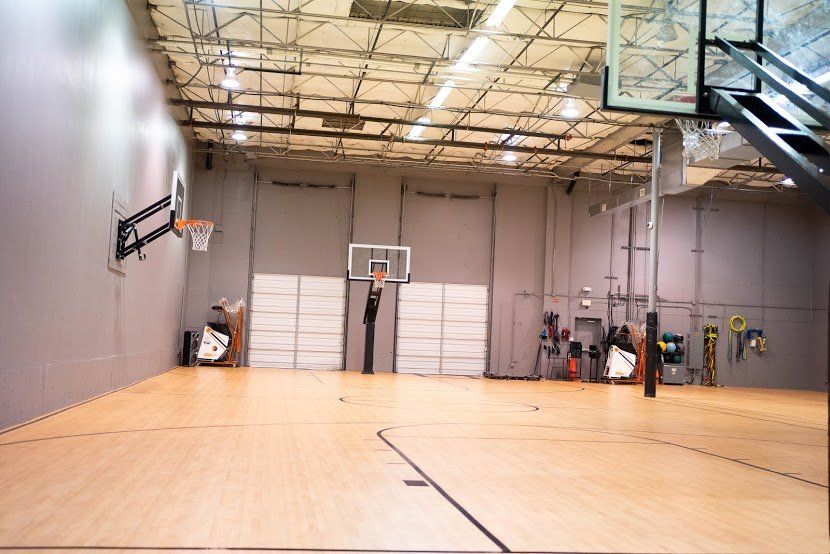 Empty indoor basketball court, hardwood floor, three hoops, gray walls, and a high ceiling.