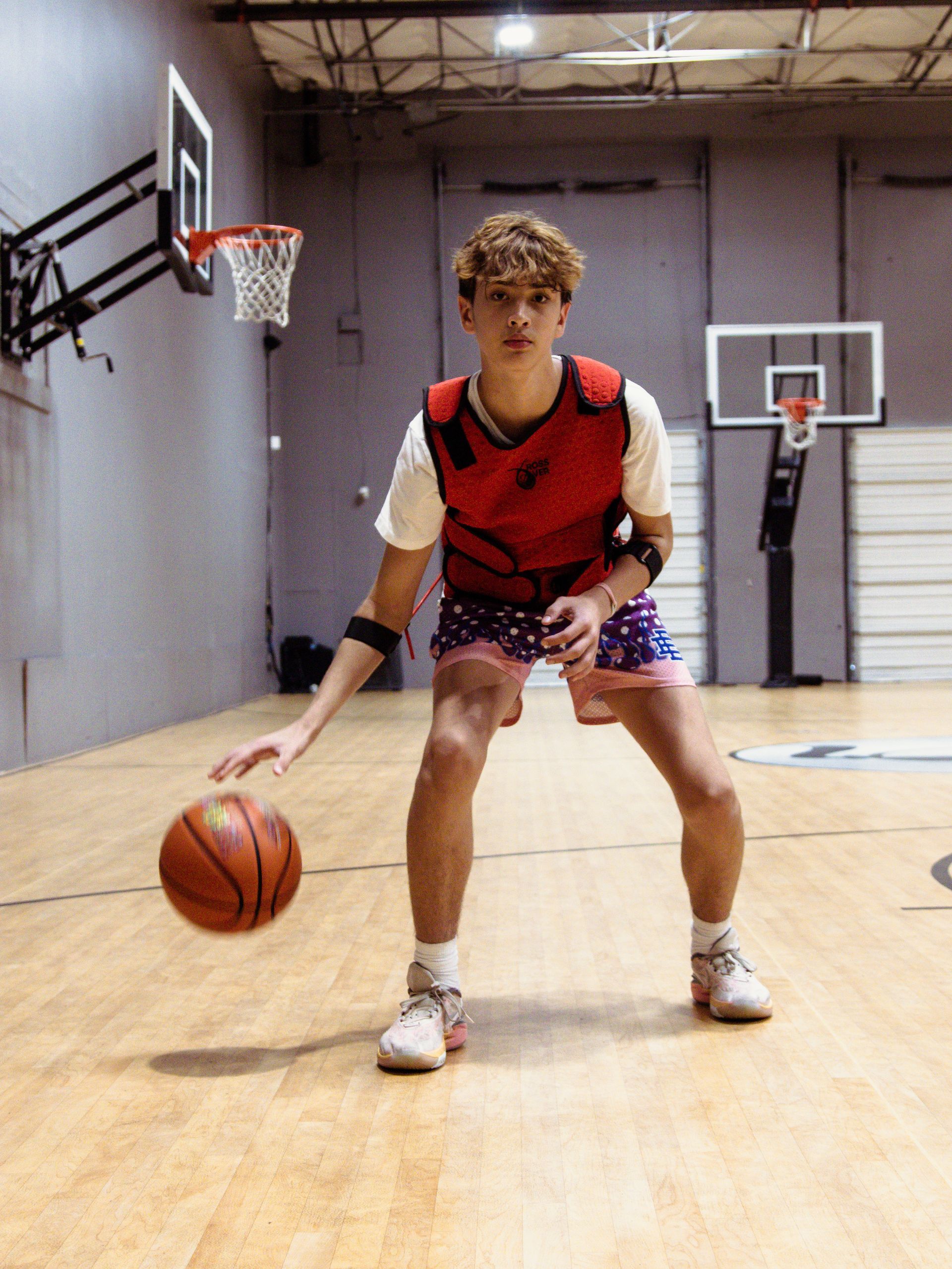 Young basketball player dribbling a ball in a gym, wearing a red vest, shorts, and sneakers.