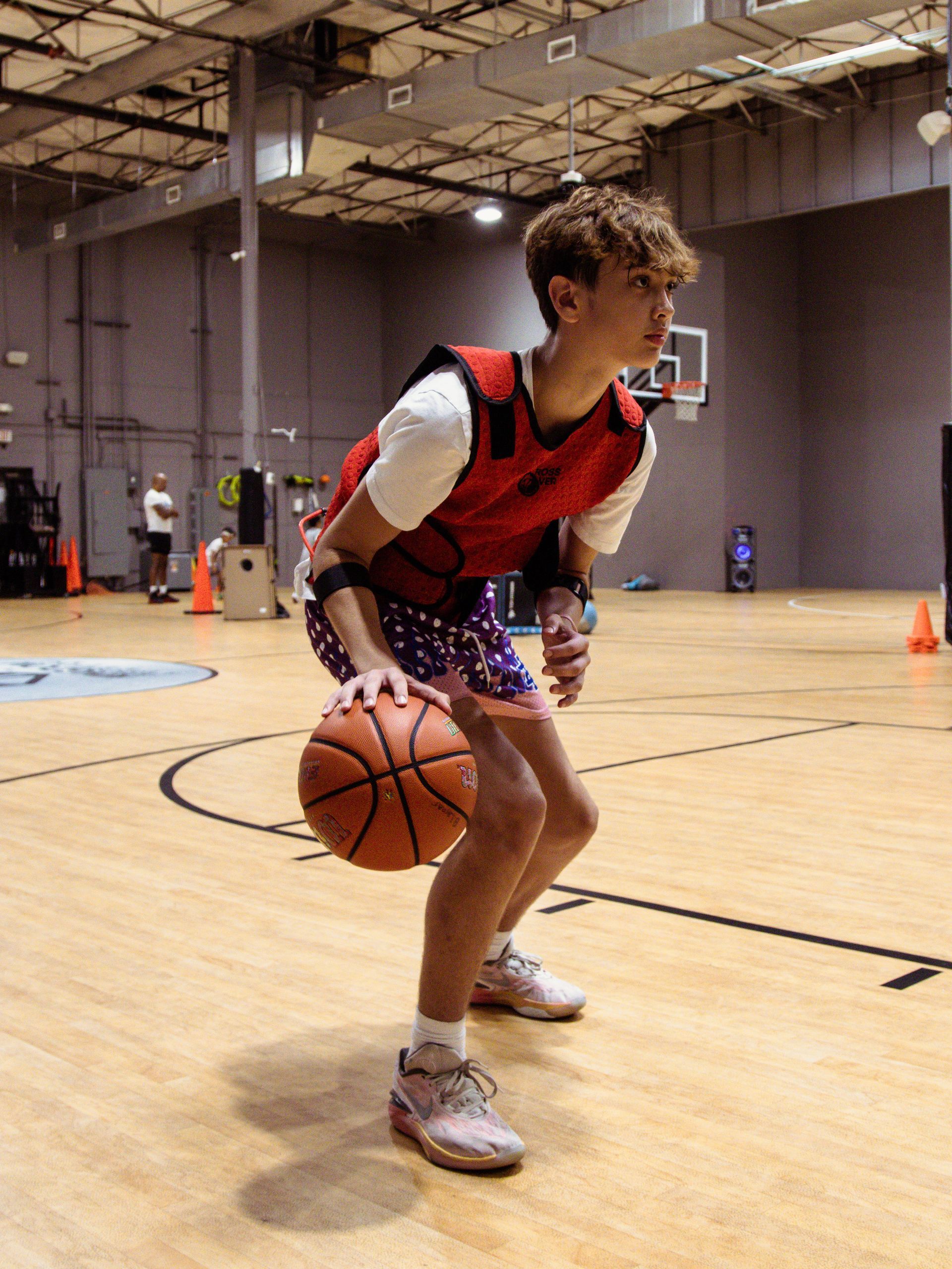 Basketball player dribbling the ball indoors, wearing a red vest and patterned shorts.