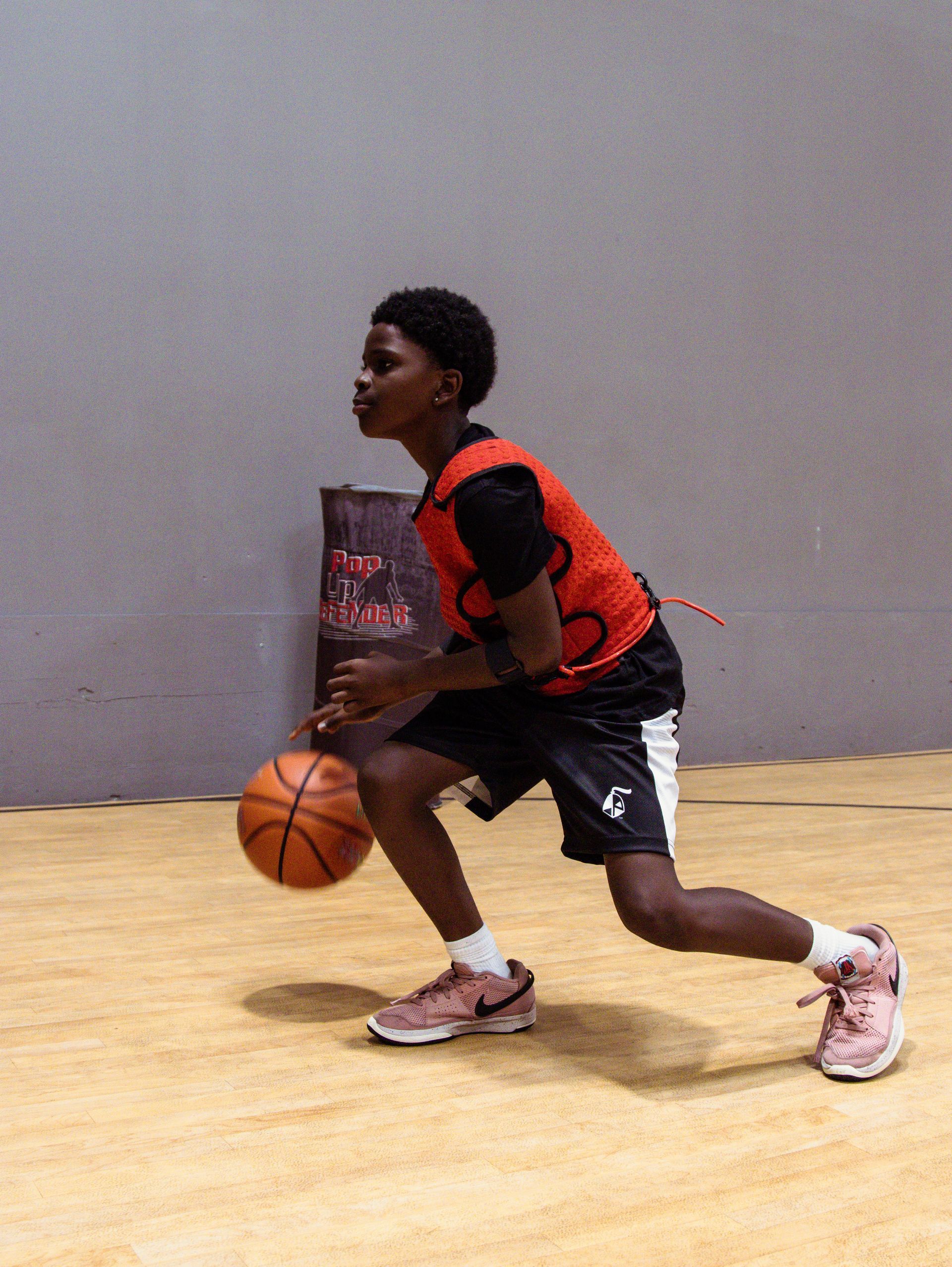 Boy in basketball stance dribbles ball indoors, wearing black shorts and orange vest.