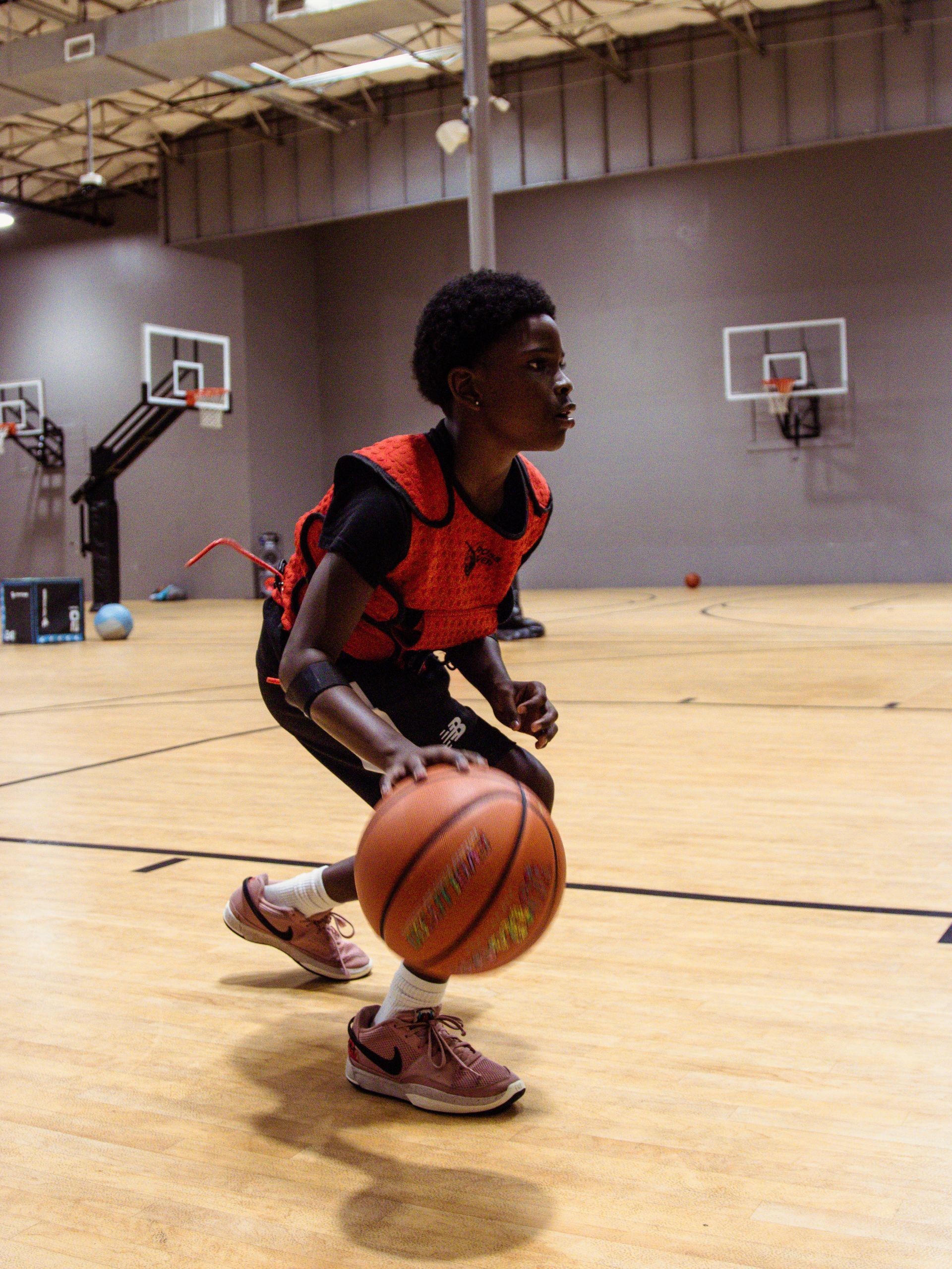 Basketball player dribbling the ball on a wooden court, wearing red vest, Nike shoes.