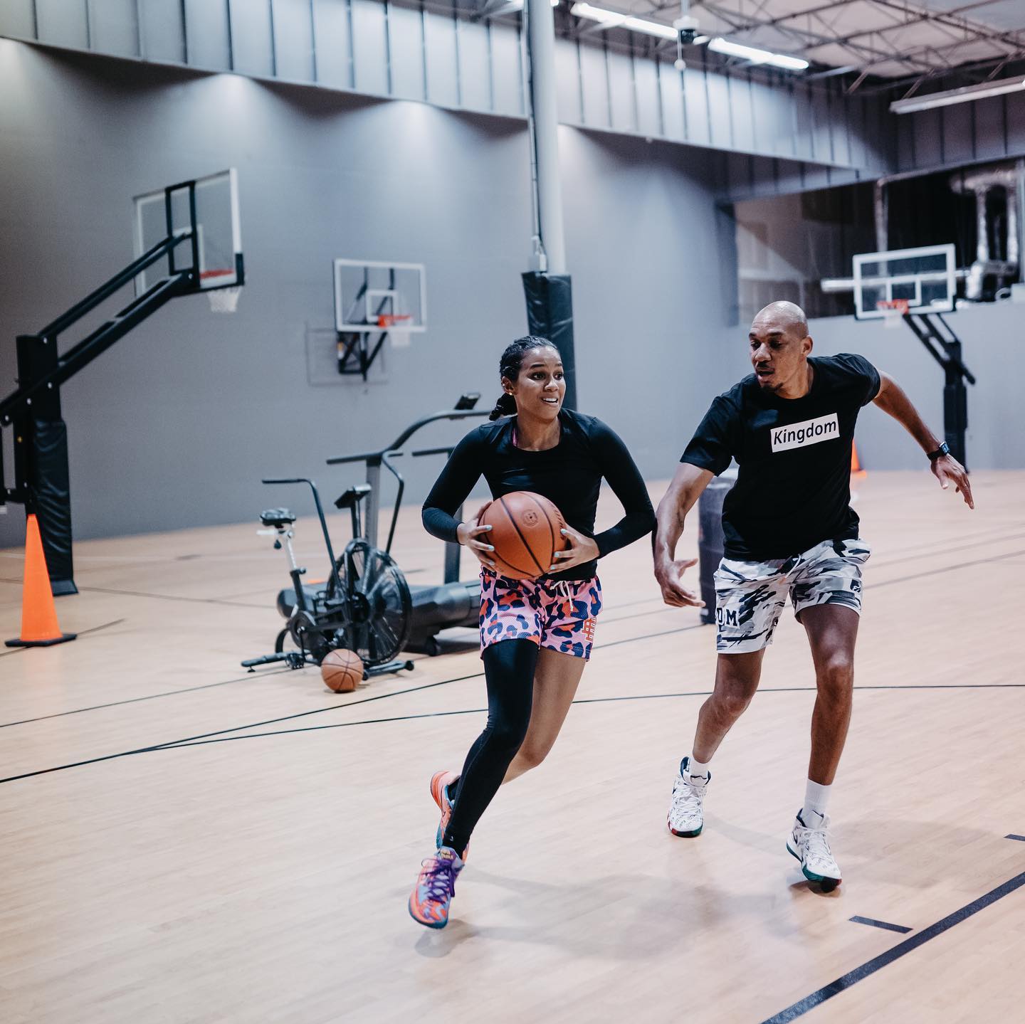 Woman dribbling basketball, chased by a man in a gym; both are smiling.