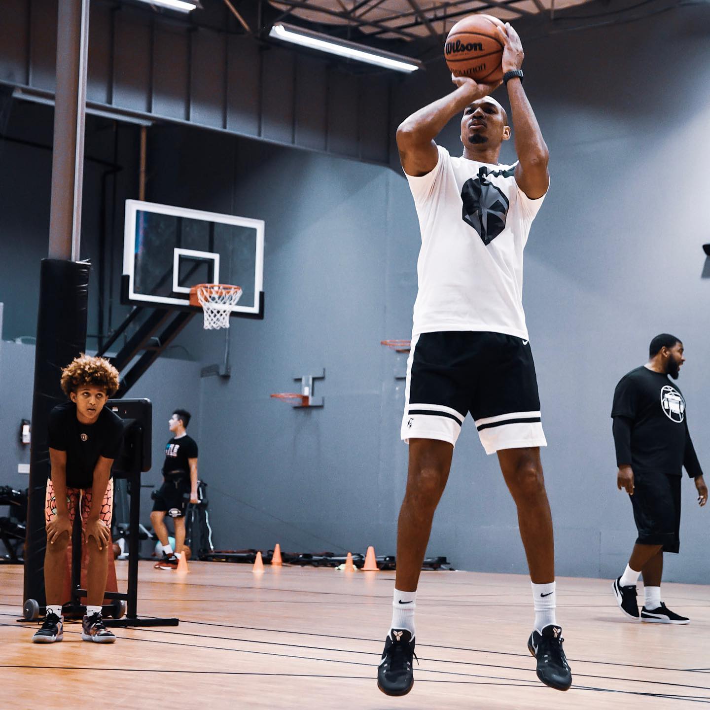 Basketball player shooting in gym, wearing white t-shirt and black shorts. Other players and equipment visible in background.