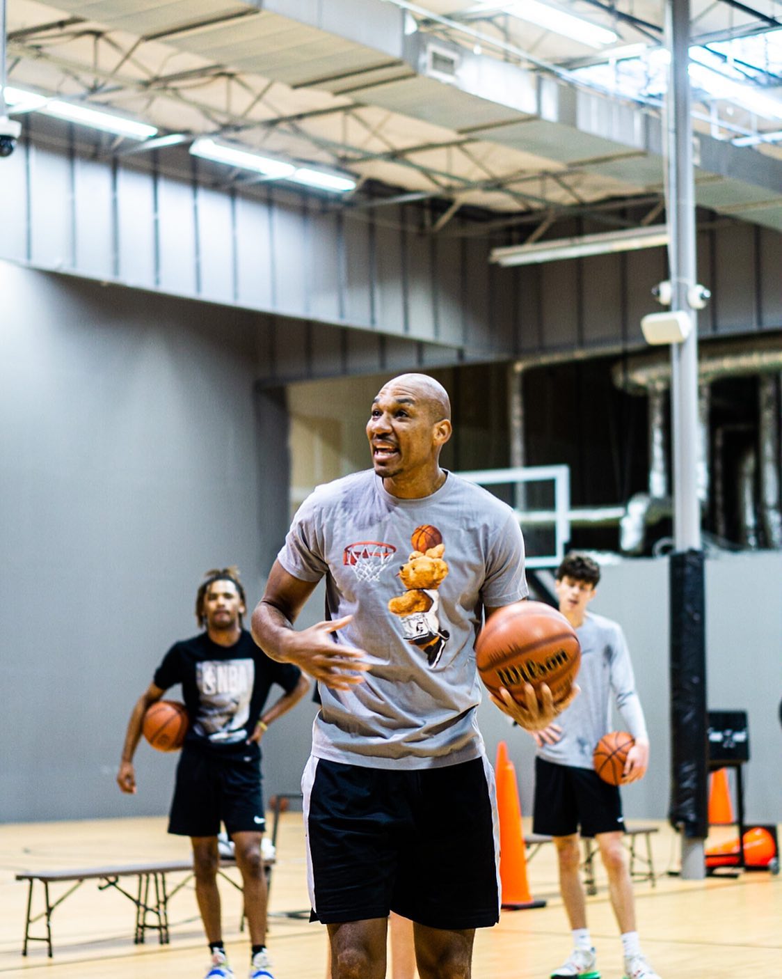 Man in a basketball gym coaching, gesturing while holding a ball, with two others holding balls behind him.