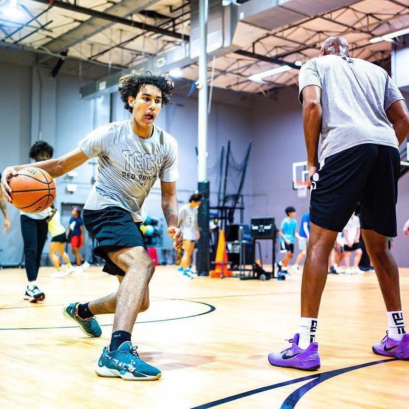 Young basketball player dribbles past a taller man, both on court. Other players in the background.