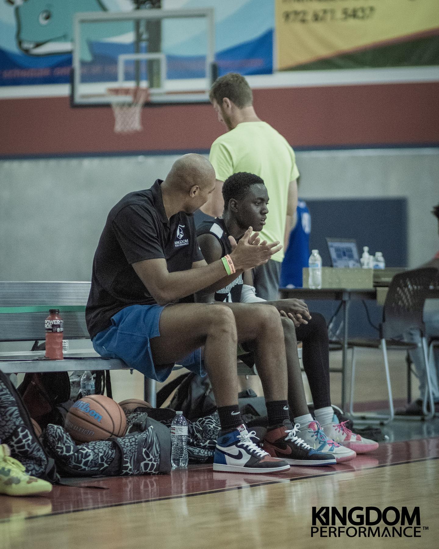 Coach and player sitting on bench, discussing strategy during a basketball game. Indoor court, bright lighting.