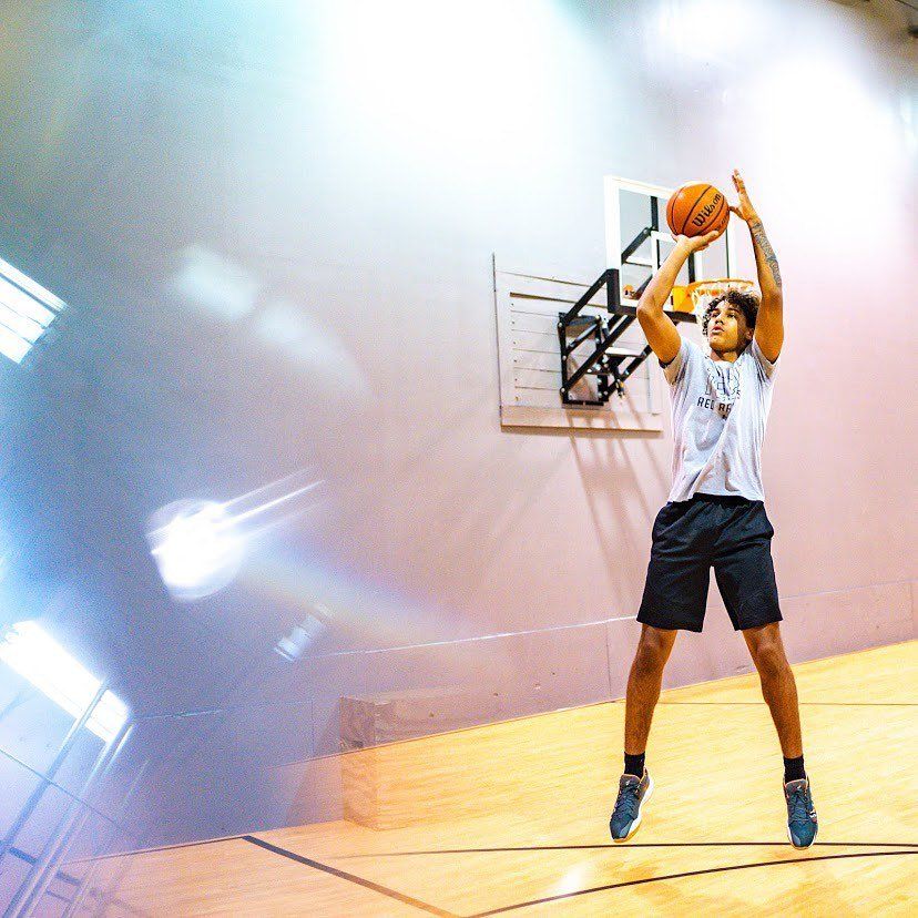 A young man shoots a basketball in a gym, leaping towards the hoop.