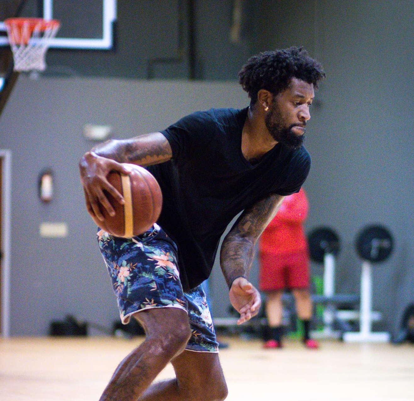 Man dribbling a basketball in a gym; wearing black shirt and floral shorts.