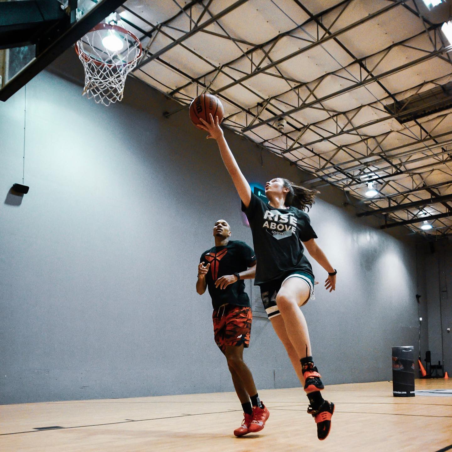 Woman in black uniform shoots a basketball in an indoor gym, man running behind.