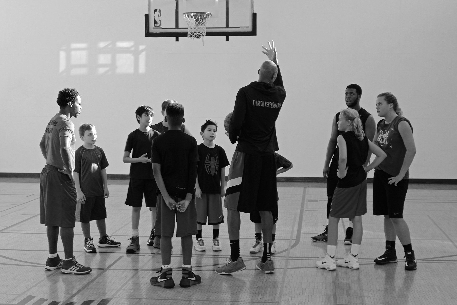 Basketball coach demonstrating a shot to a group of young players in a gym.